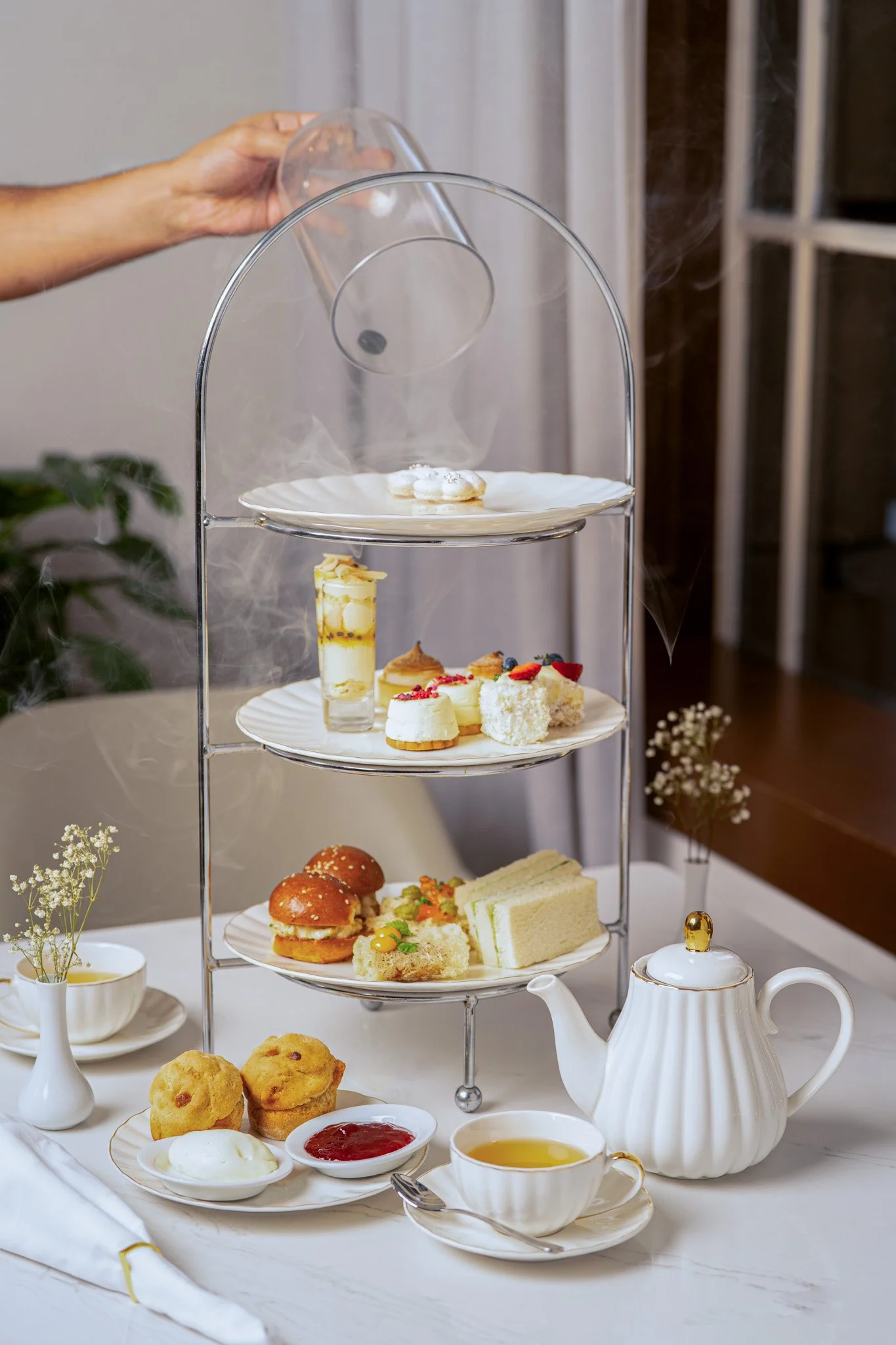 A three-tiered silver and white serving stand holding assorted desserts, sandwiches, and snacks at a tea party, with a white teapot, cups, and small bowls of tea, jam, and cream on a white tablecloth.