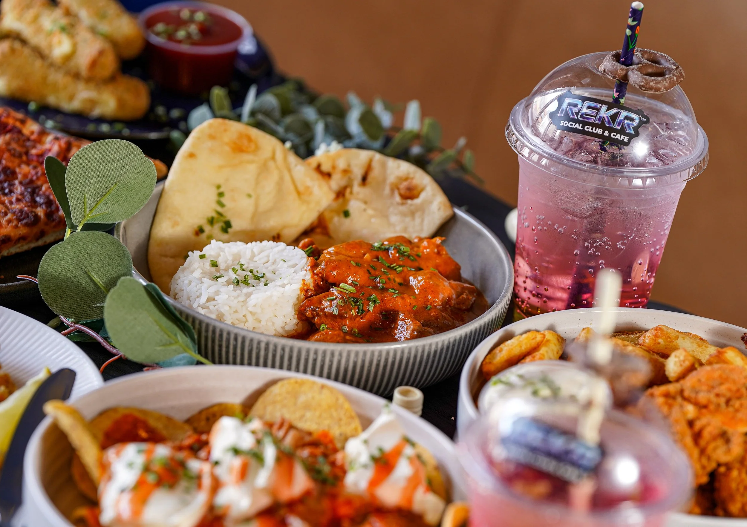 A tray of Indian cuisine dishes including rice, naan bread, curry, and fried snacks, with a pink soda in a plastic cup with a straw and a chocolate-covered pretzel on top.