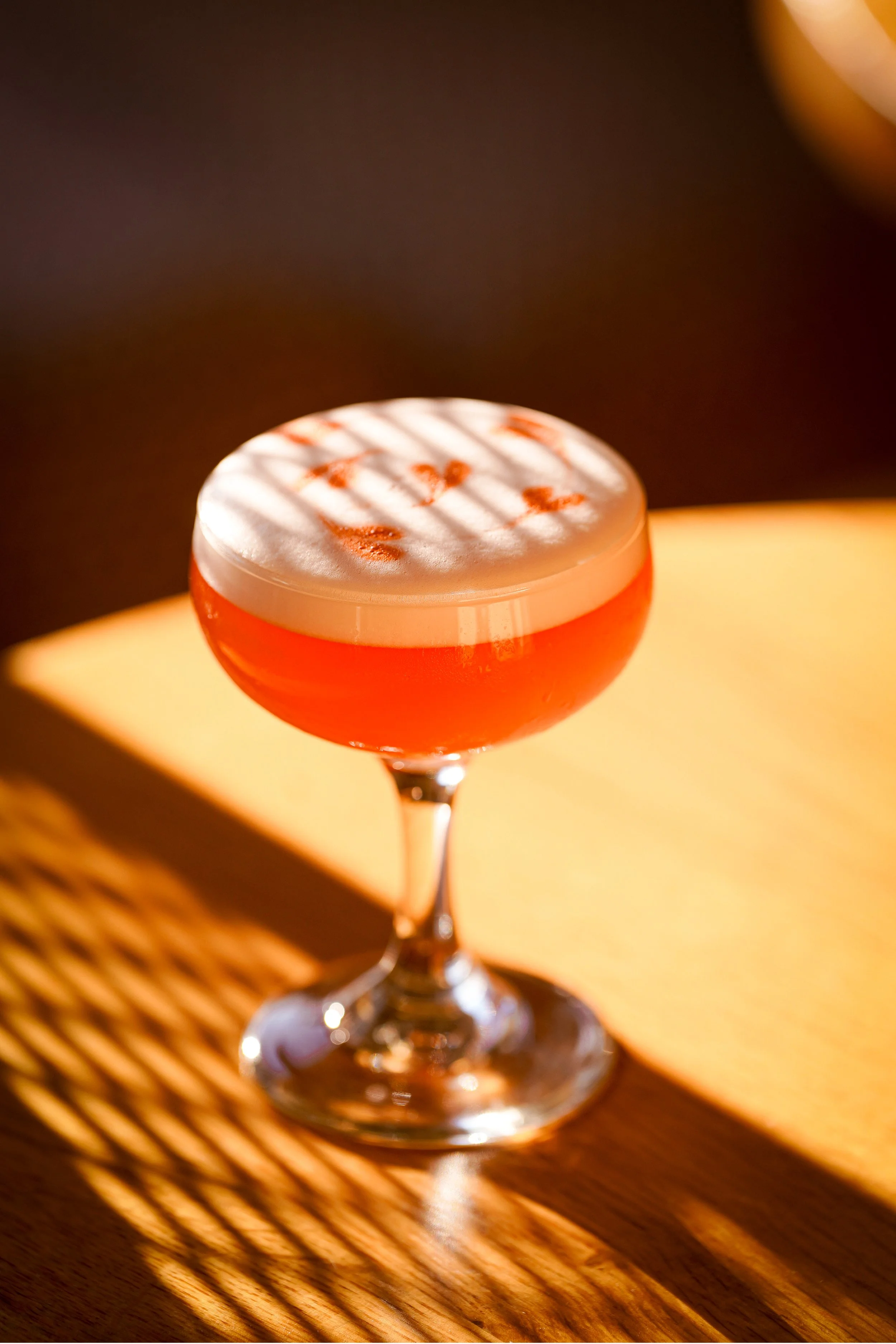 A cocktail glass filled with an orange-colored drink, topped with a frothy white layer, and garnished with red sprinkles. The glass is on a wooden surface with shadows cast by nearby blinds, shot in a restaurant in Avalon