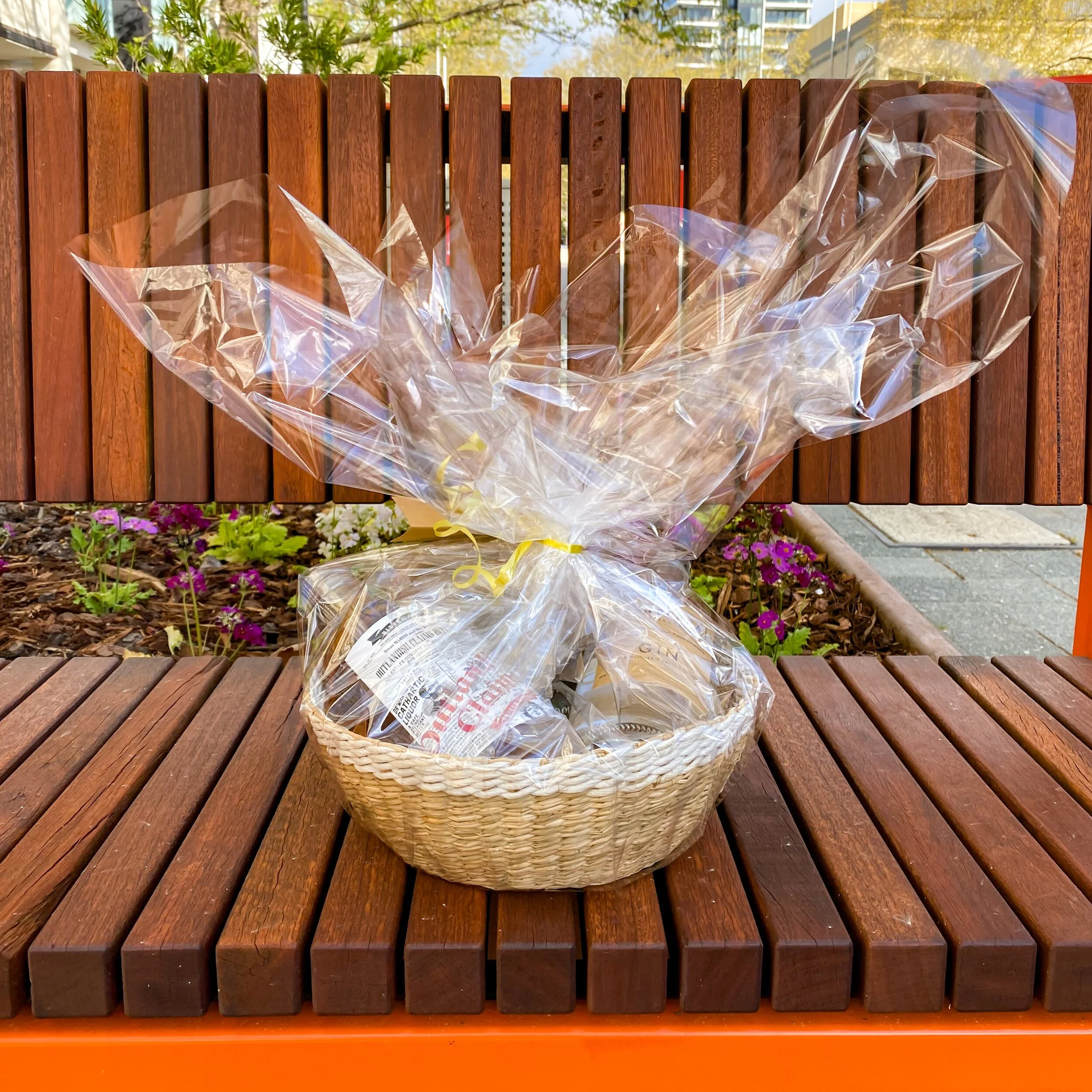 A gift basket wrapped in clear cellophane and tied with a yellow ribbon, placed on a wooden park bench with a background of flowers and plants.