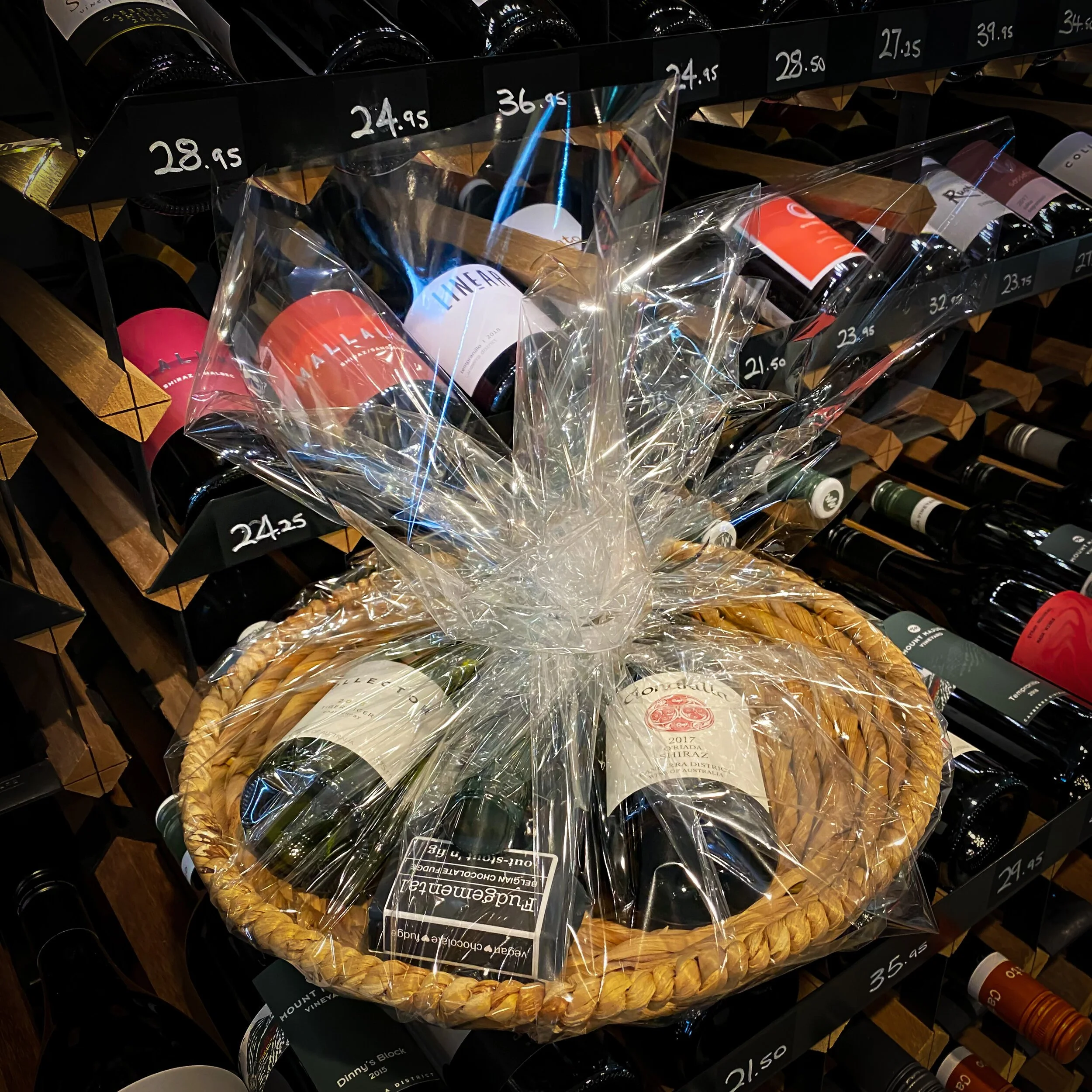 Wine bottles wrapped in clear cellophane in a woven basket on a store shelf with wine prices displayed above.
