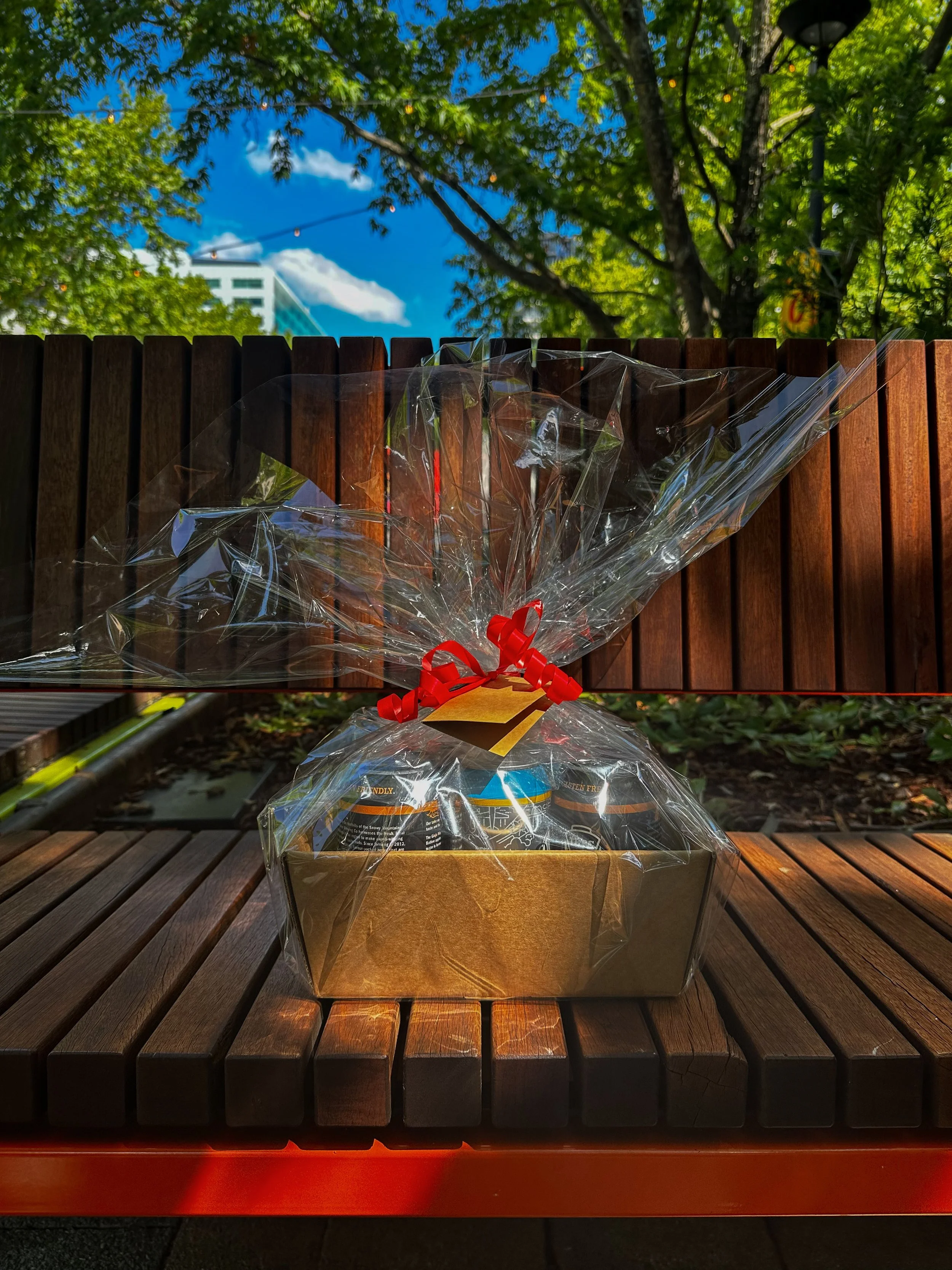 Gift basket wrapped in clear plastic with a red ribbon, placed on a wooden park bench outdoors. Trees and a building are visible in the background.