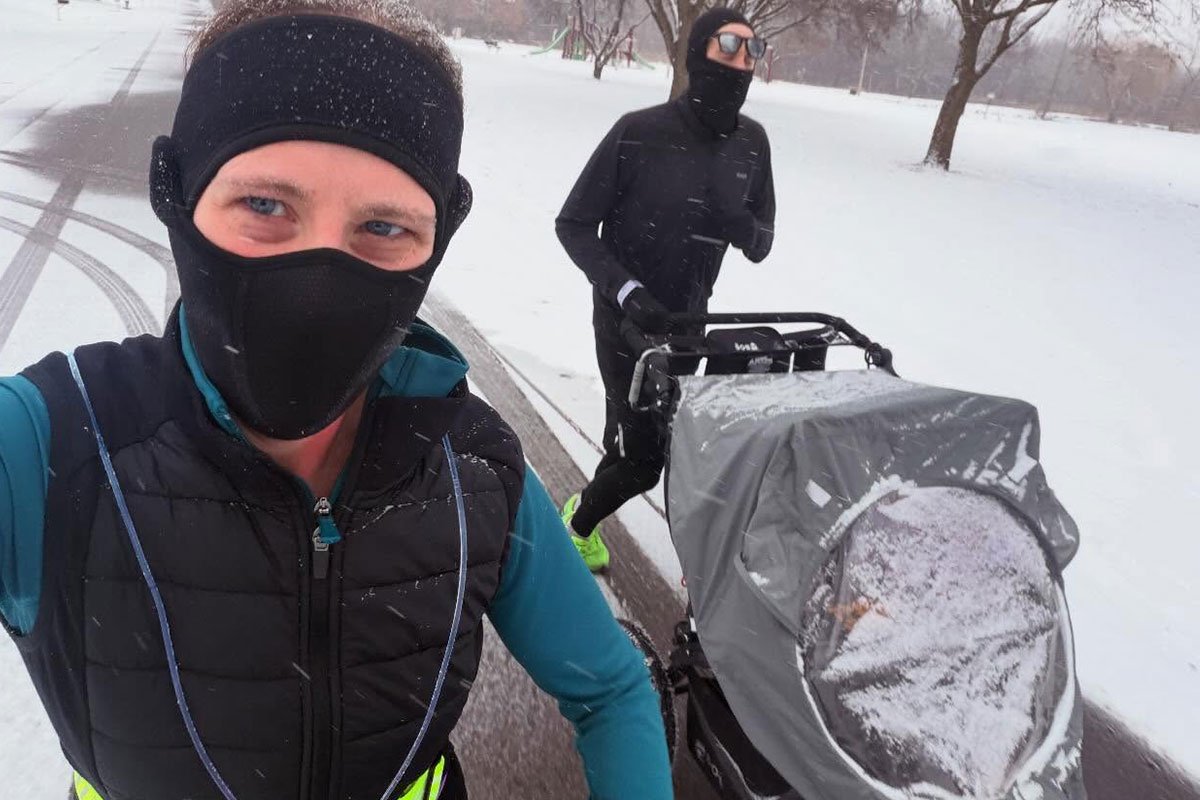 Someone in warm winter gear taking a selfie as they jog in the snow with another person pushing a stroller. 
