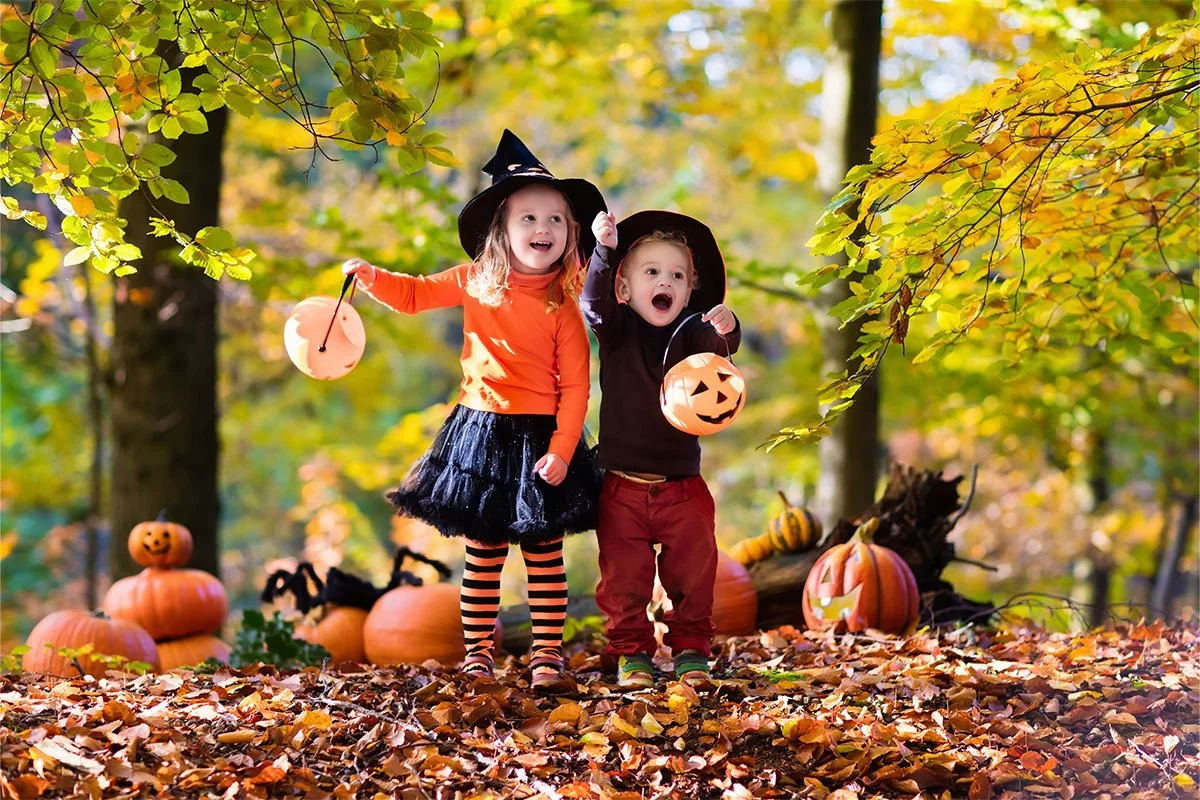Two kids dressed in halloween costumes holding up pumpkin shaped buckets and standing in fall leaves