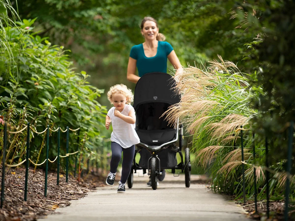 A woman running with a jogging stroller along a path outside; a child is running ahead of her. 