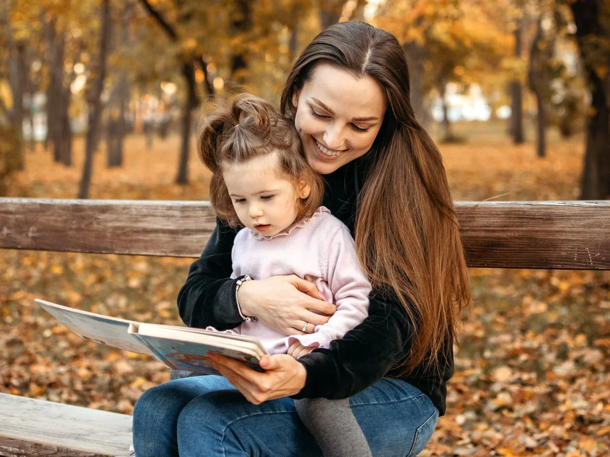 Mother with a toddler on her lap reading a book on a park bench surrounded by an autumn scenery