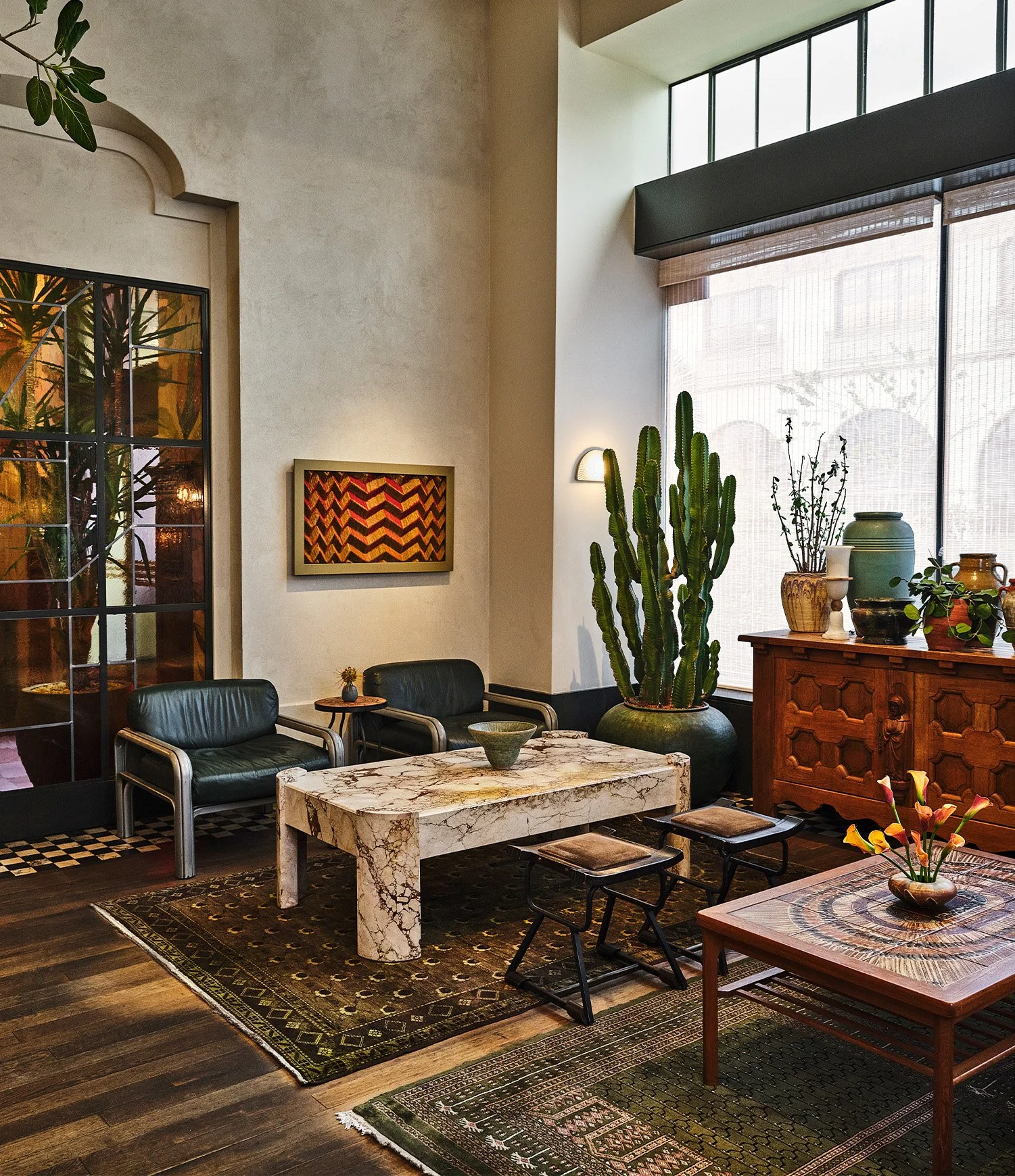 A cozy sitting area with a marble coffee table, two black leather chairs, and a side table with a small plant. Large cacti and potted plants are next to a wooden cabinet. Large windows with blinds let in natural light, and a decorative painting hangs on the wall.