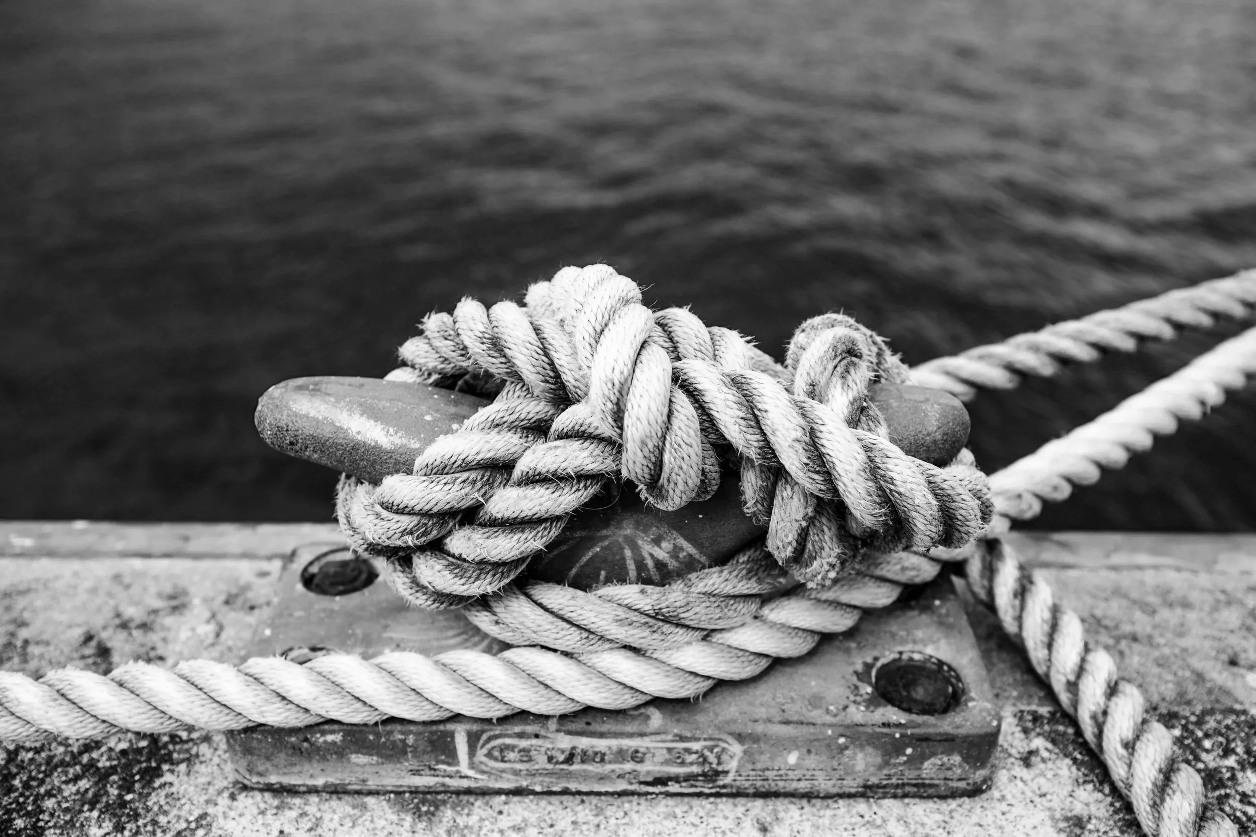 Black and white photo of a coiled rope tied around a cleat on a dock, with water in the background.