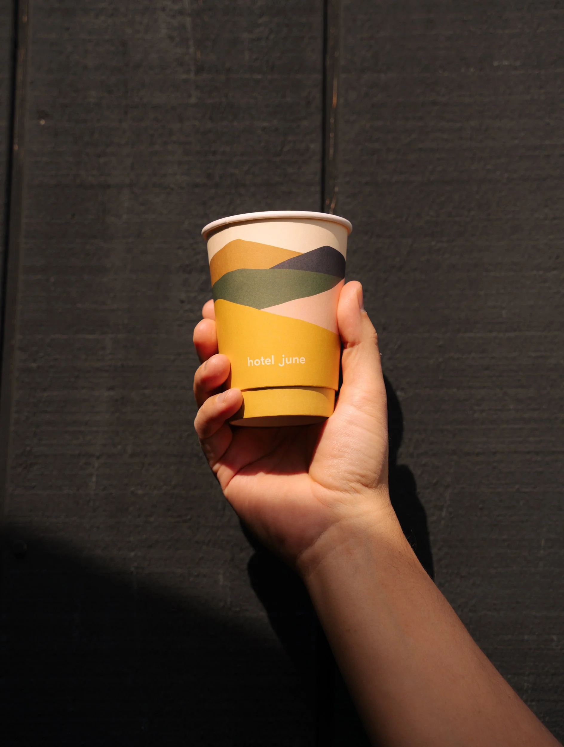 A hand holding a paper coffee cup with a mountain landscape design and the text 'hotel June' against a dark background.
