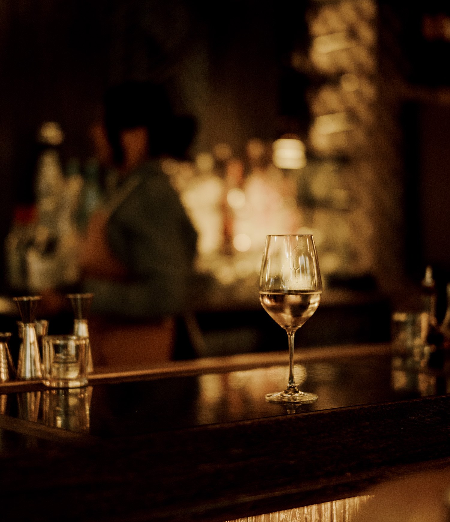 A wine glass filled with a pink-colored drink on a bar counter, with blurred bar scenery and bartender in the background.