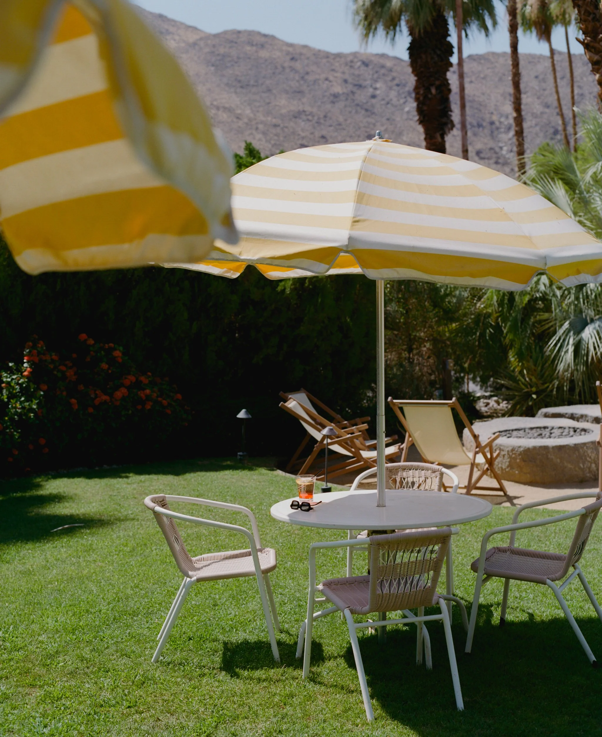 Outdoor patio area with a round white table, four white chairs, a yellow and white striped umbrella, a drink, and sunglasses on the table. In the background, there are more yellow and white striped umbrellas, lounge chairs, palm trees, greenery, and mountains.