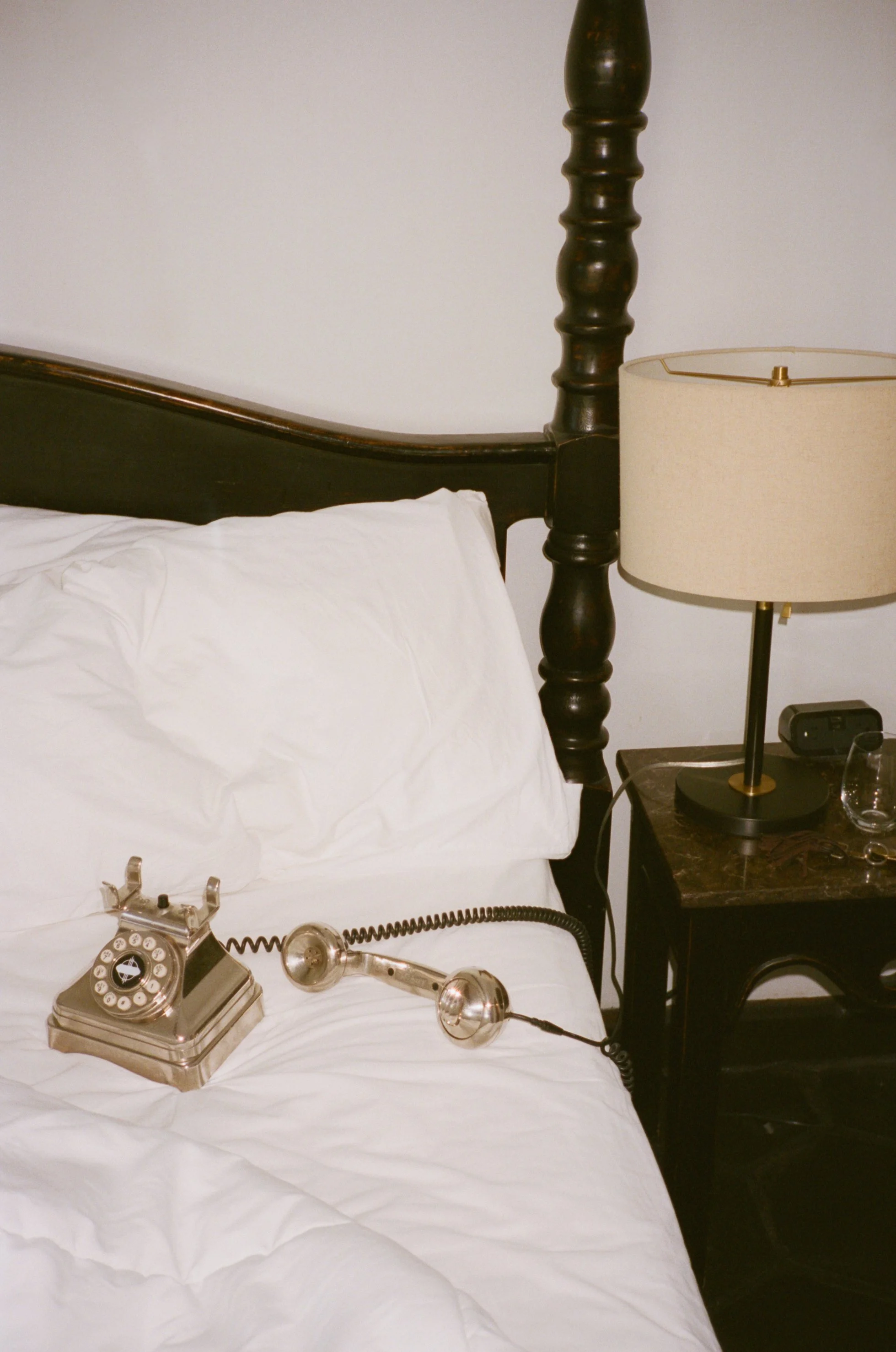 A vintage rotary dial telephone on a white bed with a black bedside table, a beige lampshade on a lamp, and a plain white wall behind.