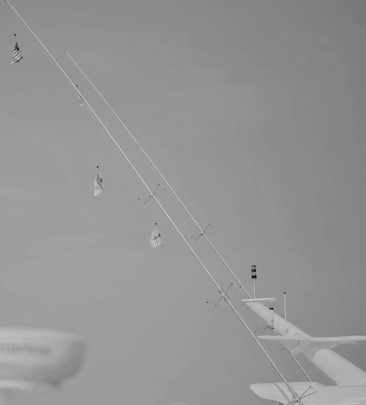 A black and white photo of a tall radio or communication tower with various antennas and flags, set against a cloudy sky, with some land or water visible at the bottom of the image.