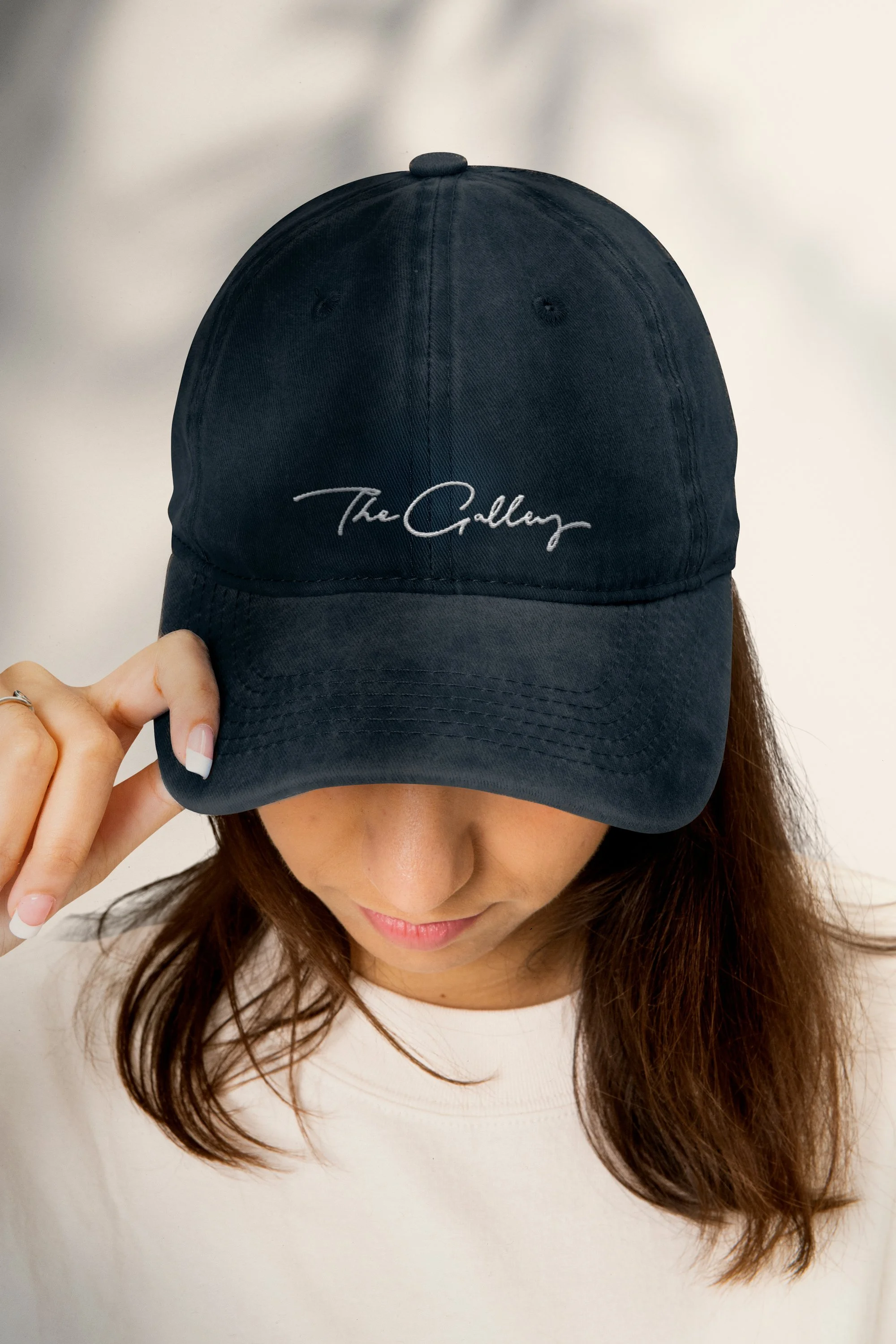 A woman with long brown hair wearing a white shirt, holding a black baseball cap with her left hand, the cap has the words 'The Callow' embroidered on it, covering part of her face and eyes.