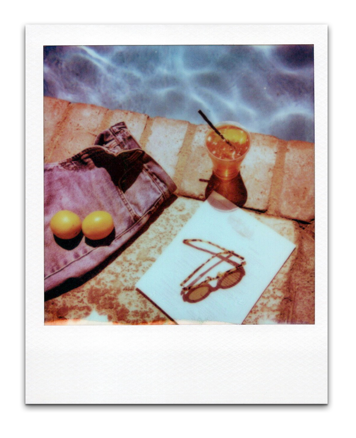 Polaroid photo of a poolside scene with a pair of jeans, two yellow lemons, a white napkin with sunglasses drawing, a glass of yellow cocktail with ice and a straw, and a white towel, against a brick ledge and blue water background.