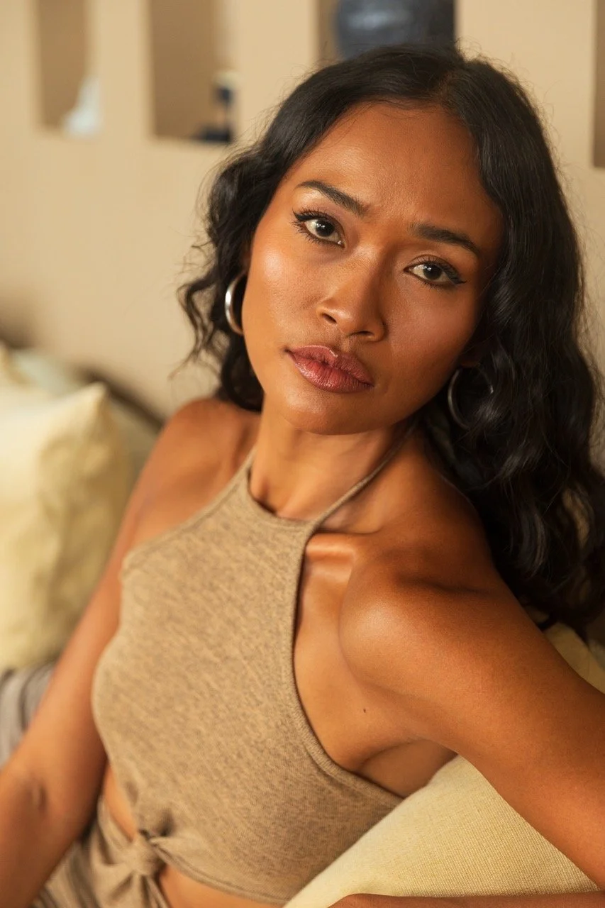 A young woman with dark wavy hair and hoop earrings sitting on a beige couch, looking at the camera with a neutral expression.