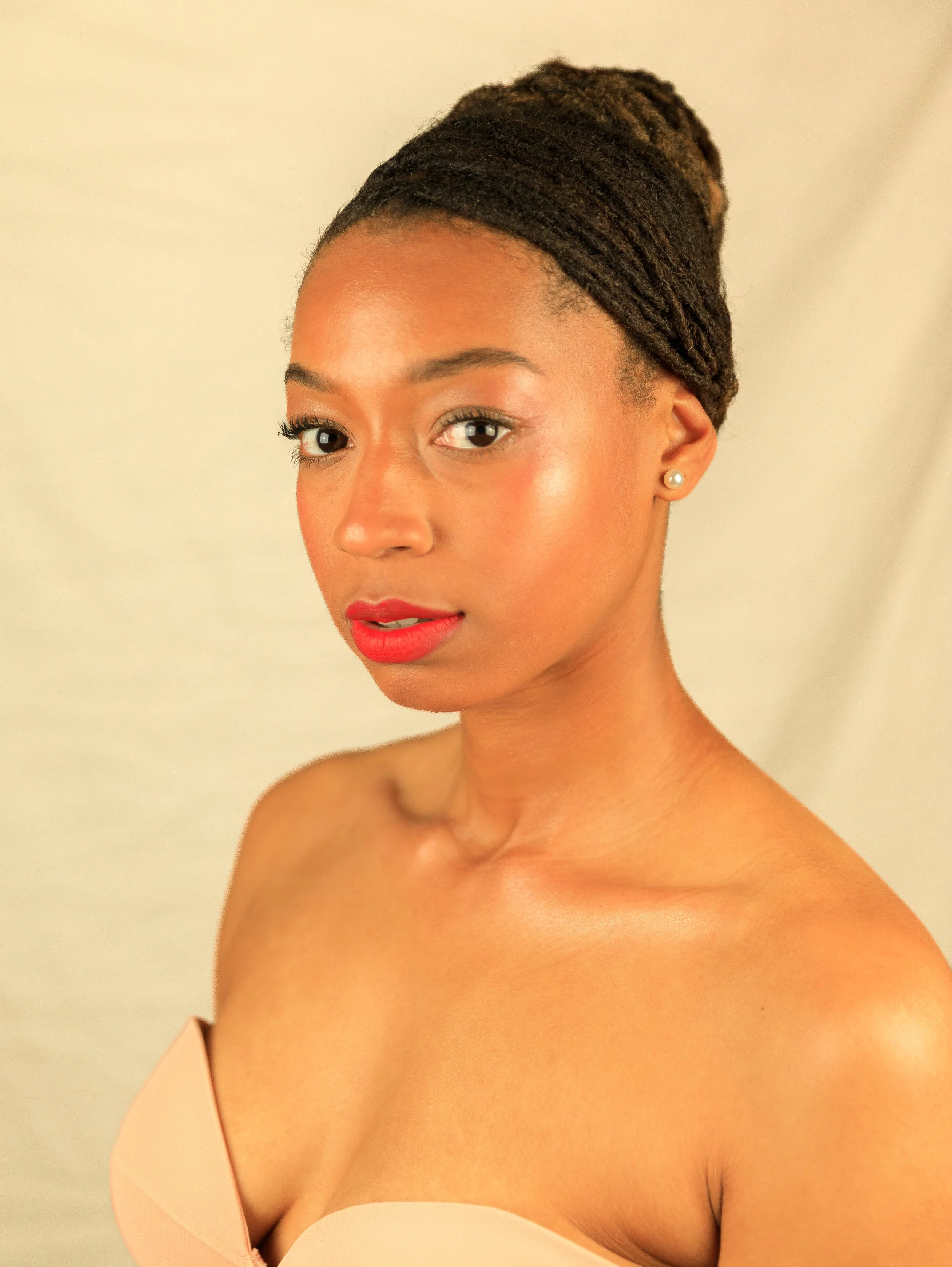 A woman with styled dreadlocks, wearing a strapless beige top and earrings, looking at the camera with a neutral expression.