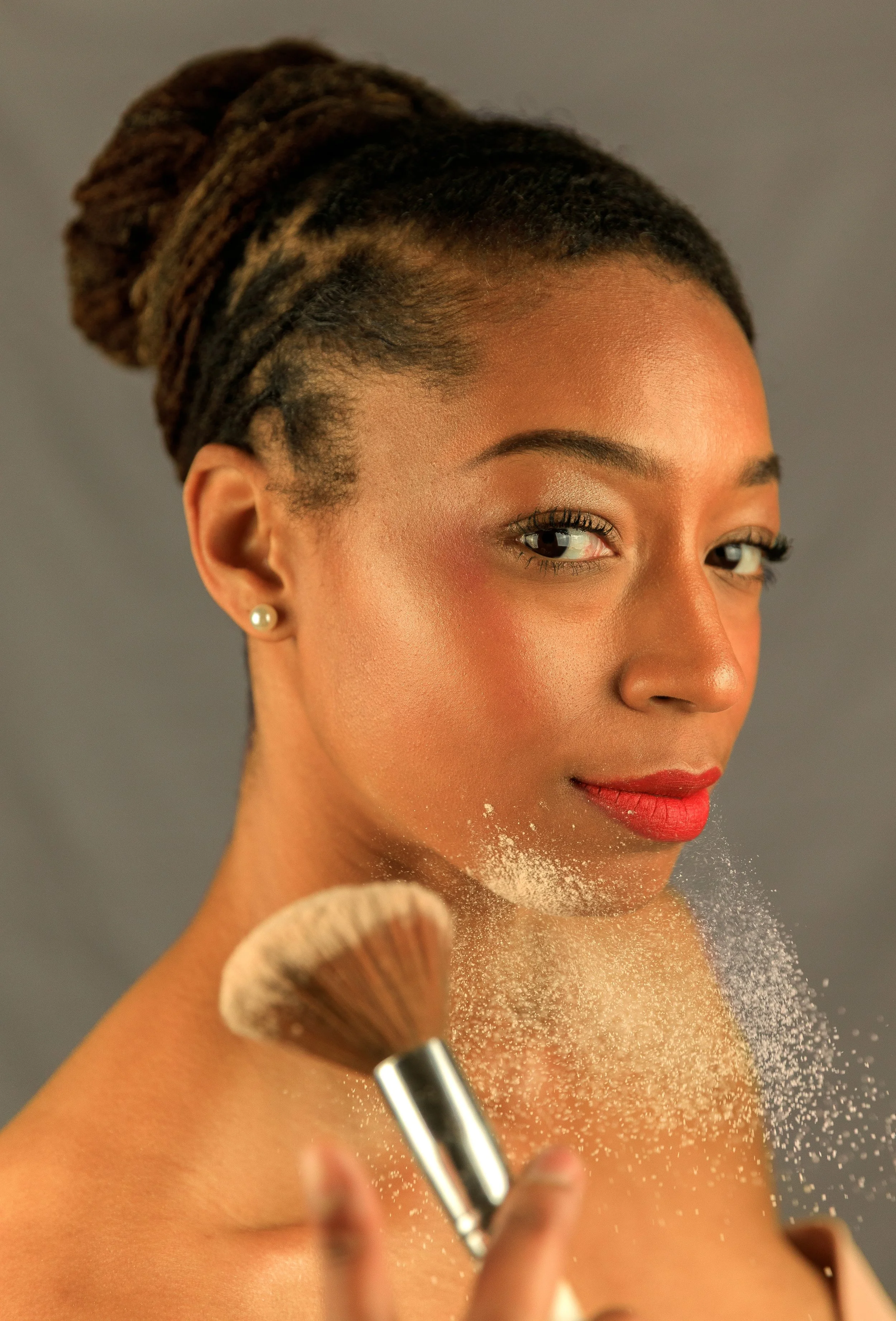A woman with braided dreadlocks, wearing pearl earrings and makeup, holds a makeup brush with powder near her shoulder.