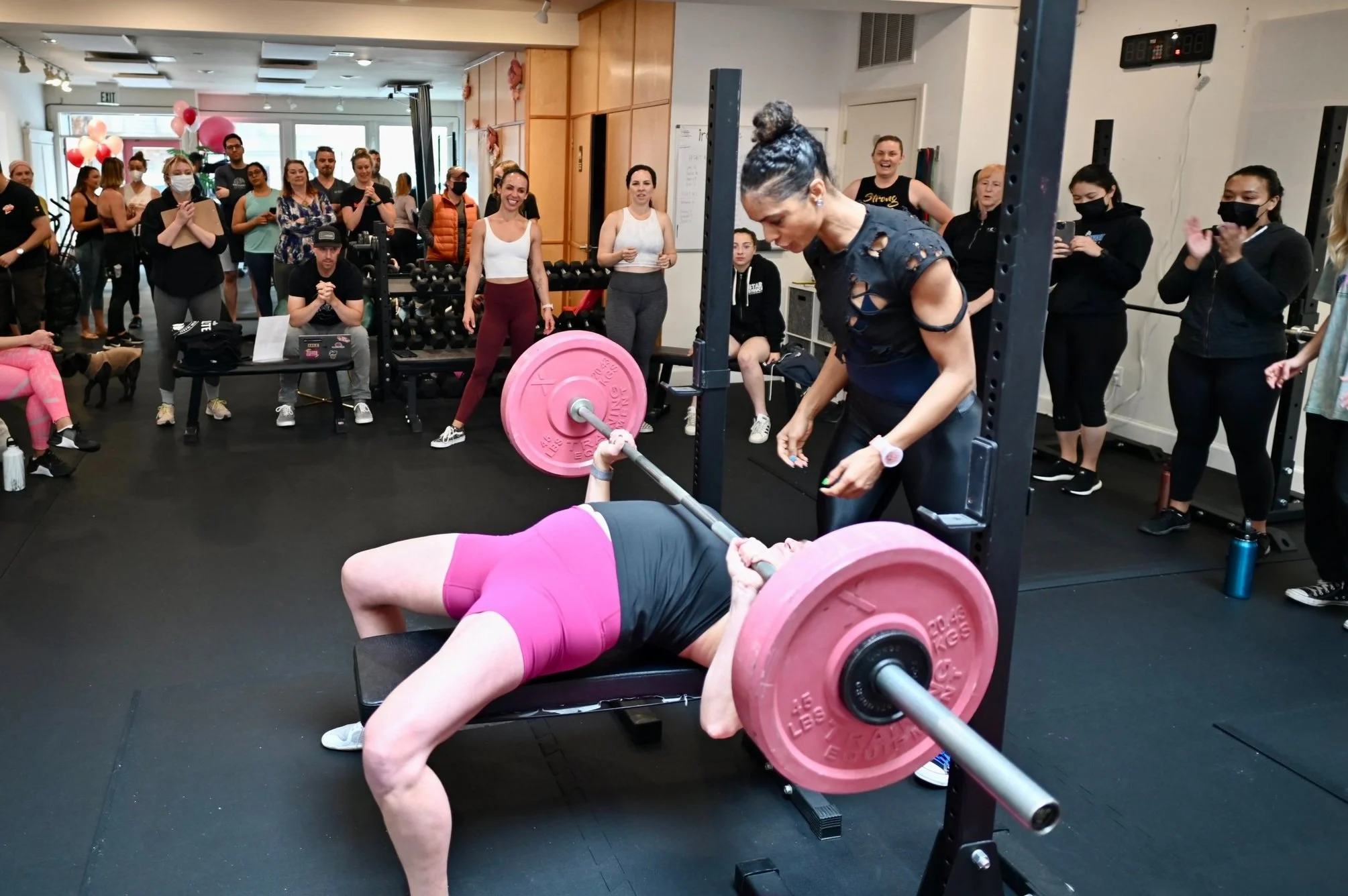 A woman in pink workout shorts and a black top performs a bench press with a pink barbell while a trainer assists. Multiple spectators observe in a gym with various workout equipment and pink decorations.