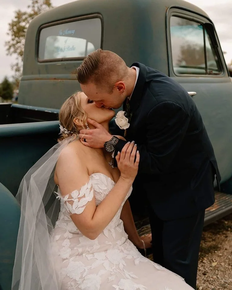 Bride and groom kissing in front of the Adler Ranch vintage truck during a summer wedding, with the bride wearing a purchased wedding dress and veil, illustrating how heirloom details like veils or headpieces can complement a modern gown.