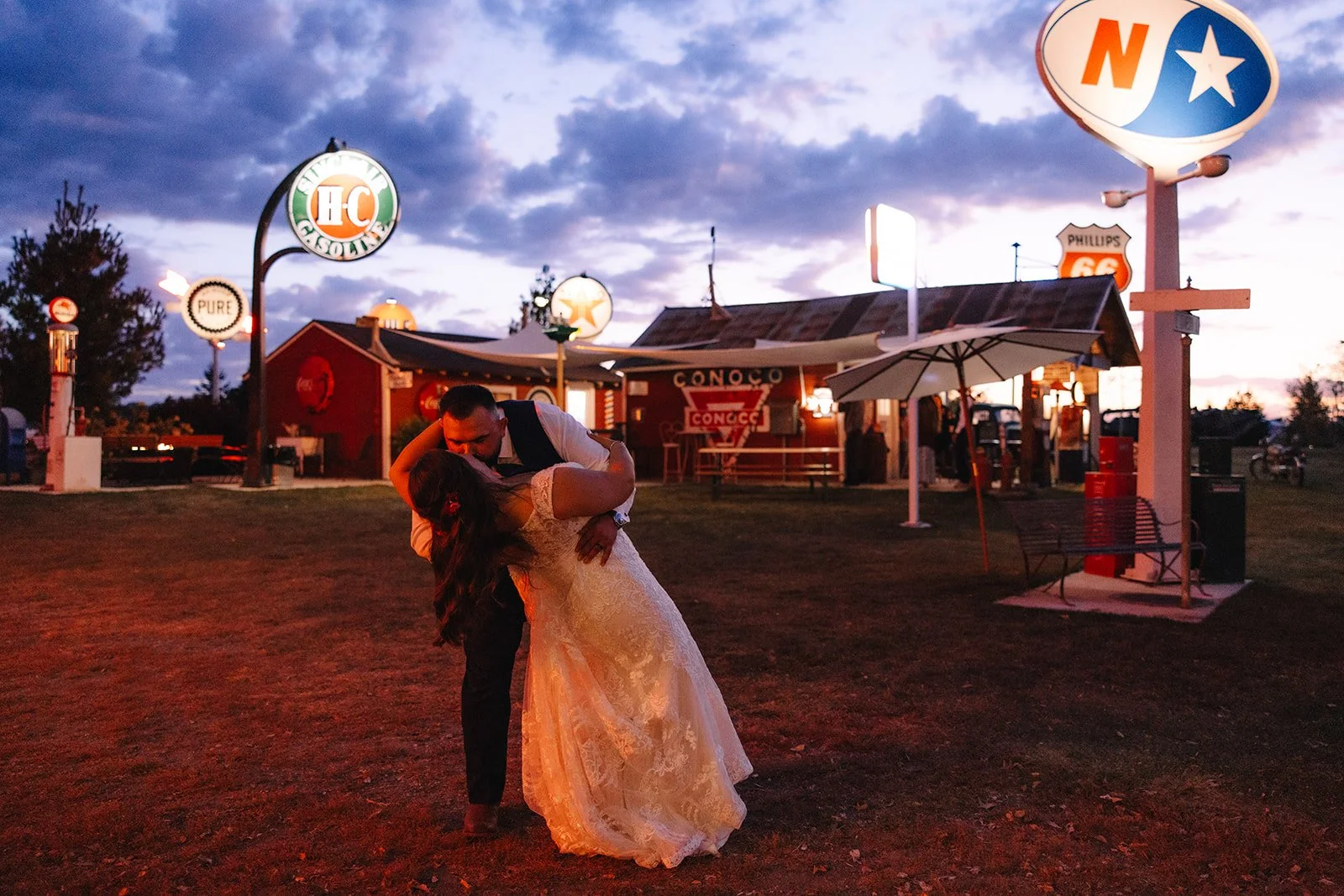 Bride and groom kissing in a dip in front of vintage gas station with neon signs at night at Adler Ranch wedding venue in Alexandria Minnesota