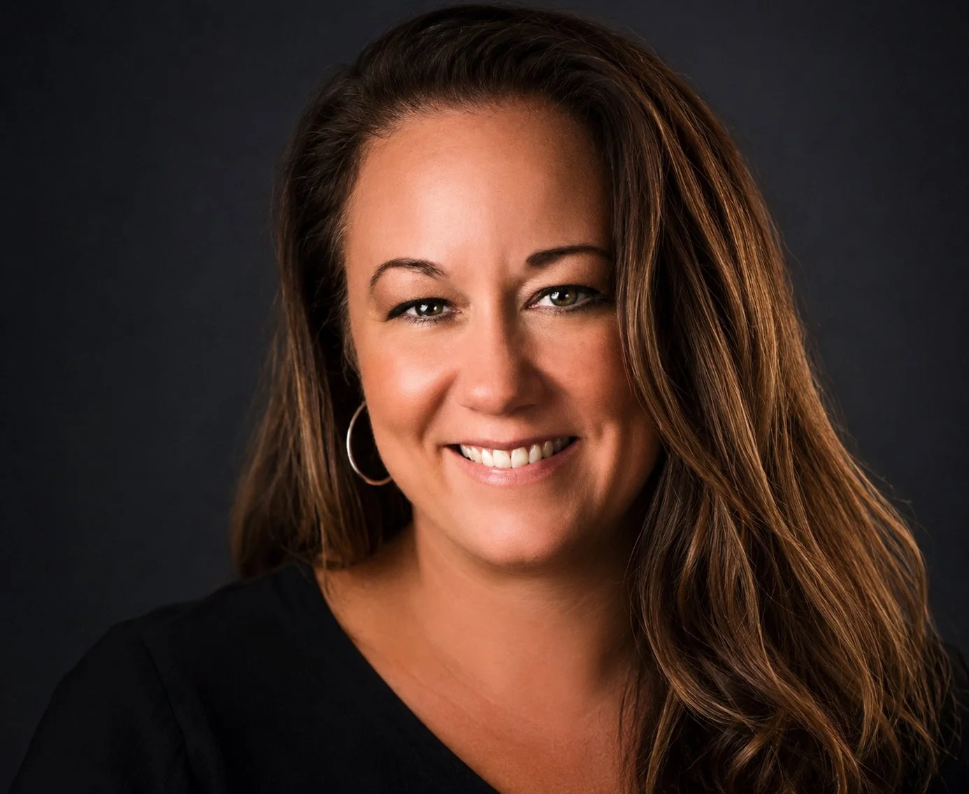 A woman with long brown hair, green eyes, wearing hoop earrings and a black top, smiling against a dark background.
