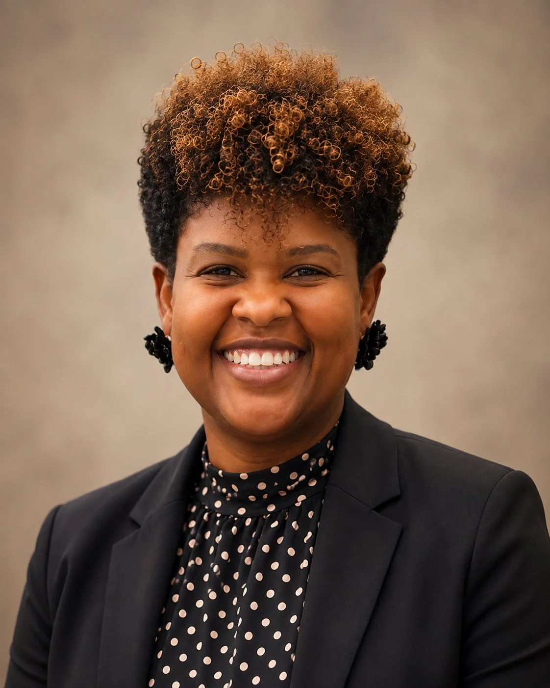 A smiling African American woman with short curly hair wearing a black blazer and a polka dot blouse.