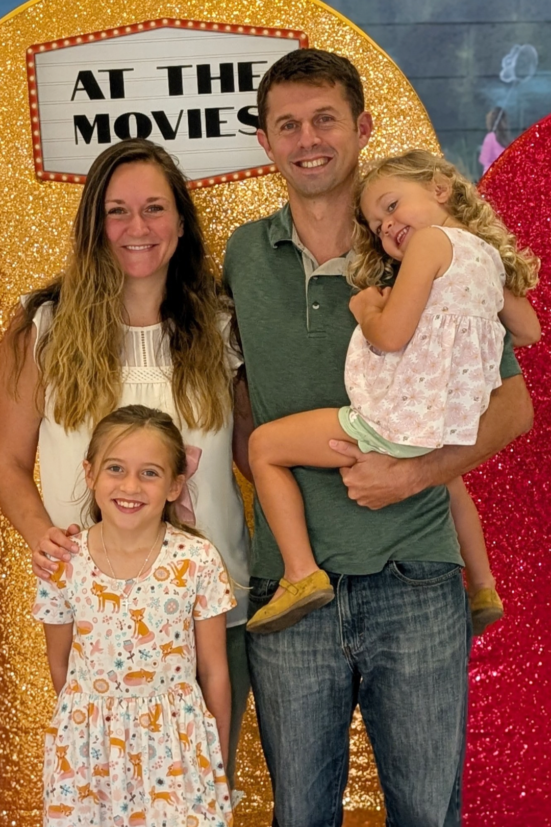 A family photo of a man, woman, and two young girls posing in front of a gold glittery backdrop with a sign that reads 'At the Movies.' The man is holding one of the girls, who is smiling and leaning on him. The woman is standing next to him with her arm around the other girl, all smiling.