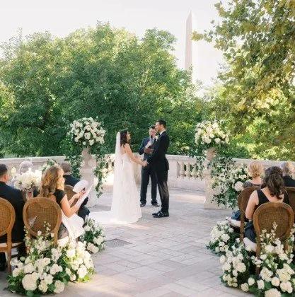 Outdoor wedding ceremony at the DAR in Washington, DC, with lush white floral arrangements framing the couple and the Washington Monument visible in the background.