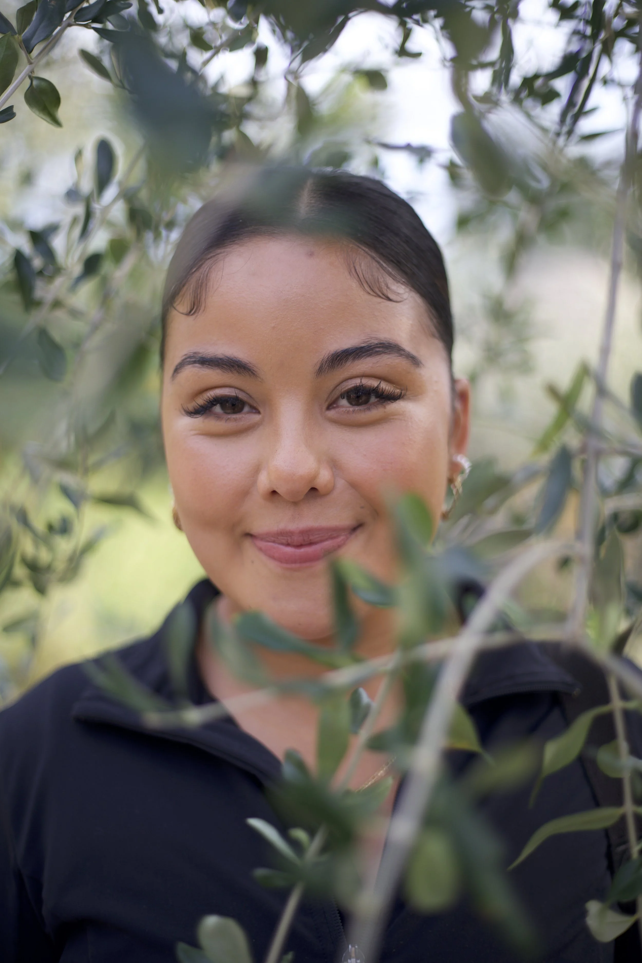 A woman with dark hair pulled back smiling amidst green foliage.