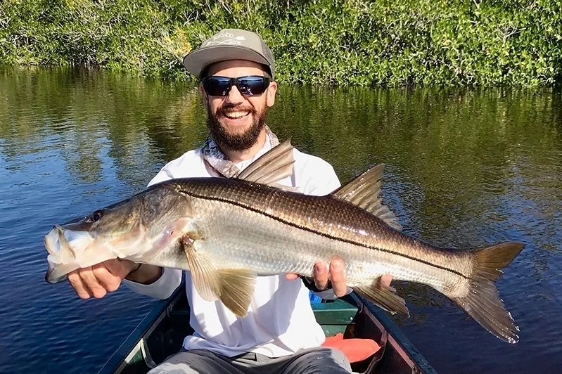 Man smiling on a boat holding a large fish with water and greenery in the background.