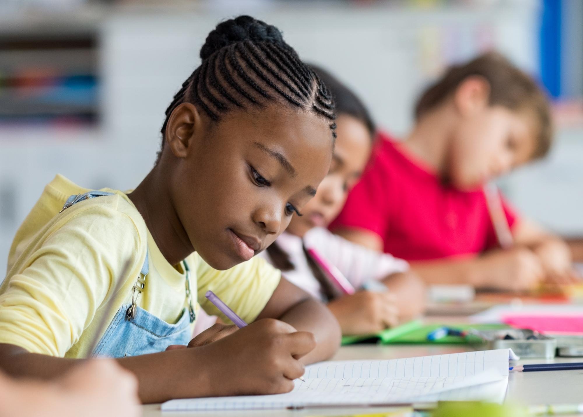 Young students sitting at a desk in a classroom, focused on writing in notebooks.