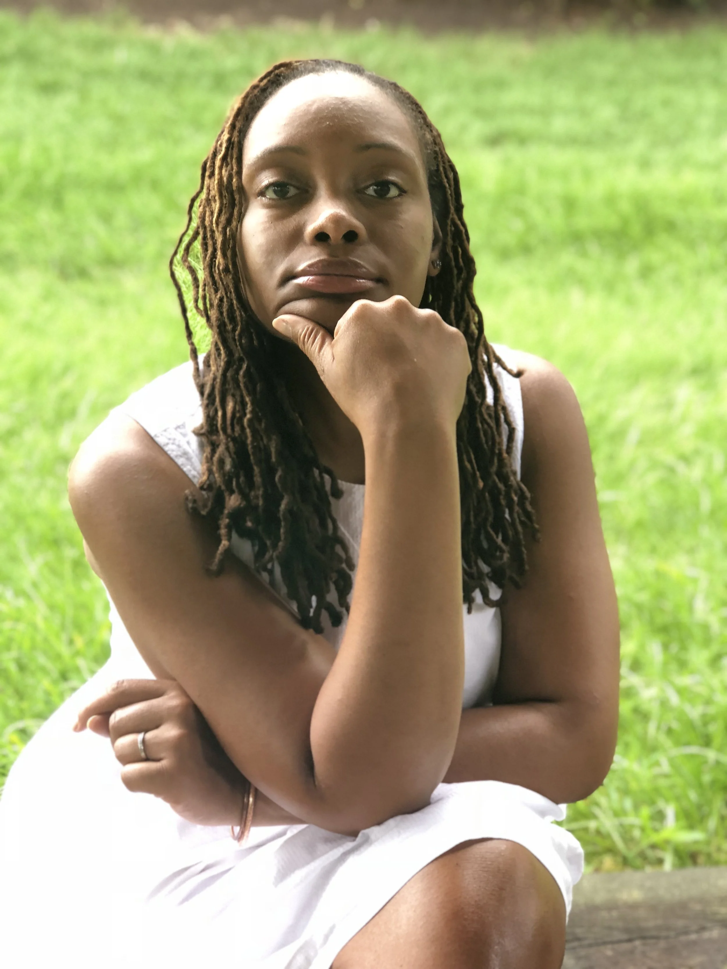 Woman with long, twisted hair, wearing a white sleeveless top, sitting outdoors on grass, resting her chin on her hand, looking contemplative.