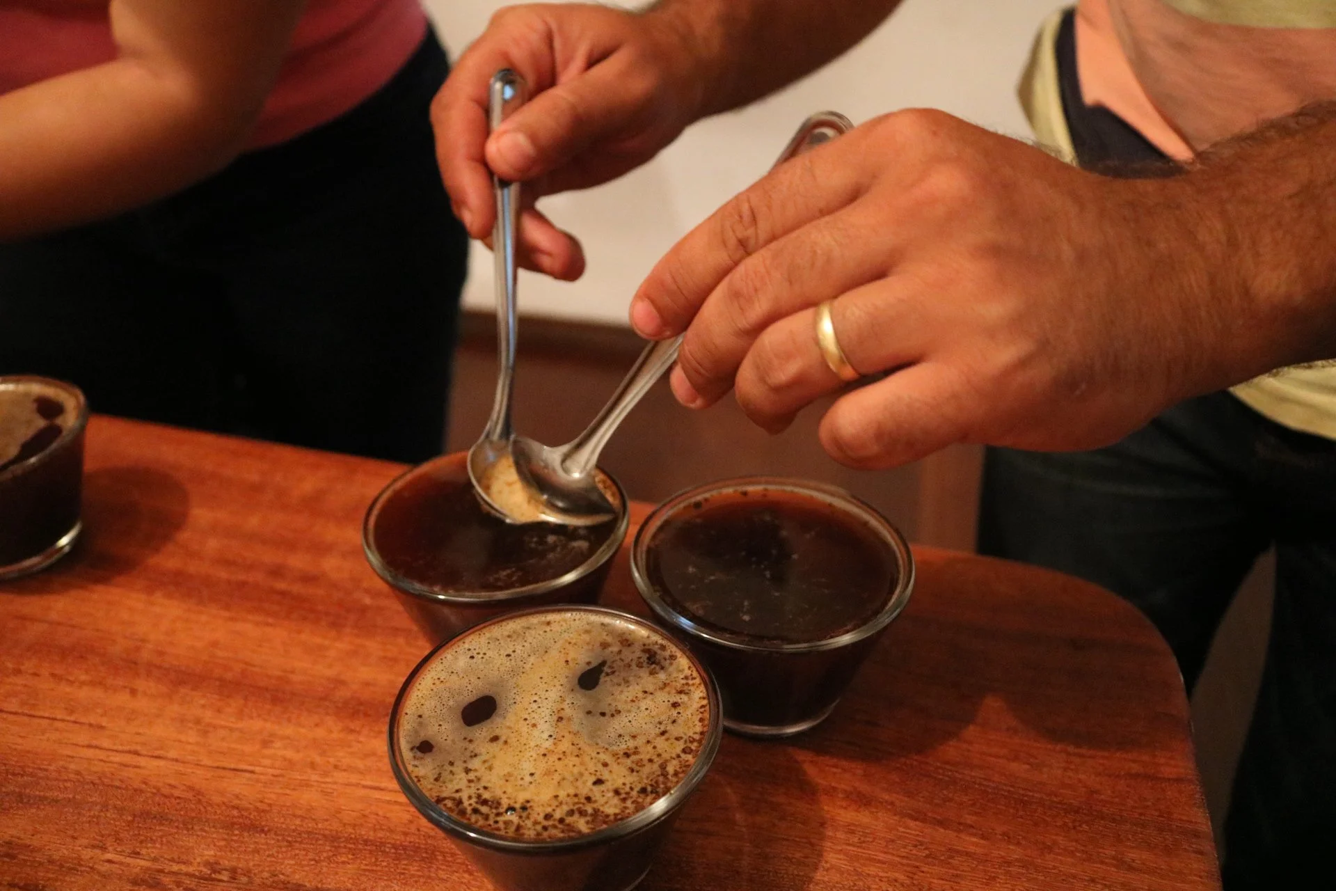 A person with a wedding ring scooping foam from a glass of dark beer, with two other glasses of similar beer on a wooden table.