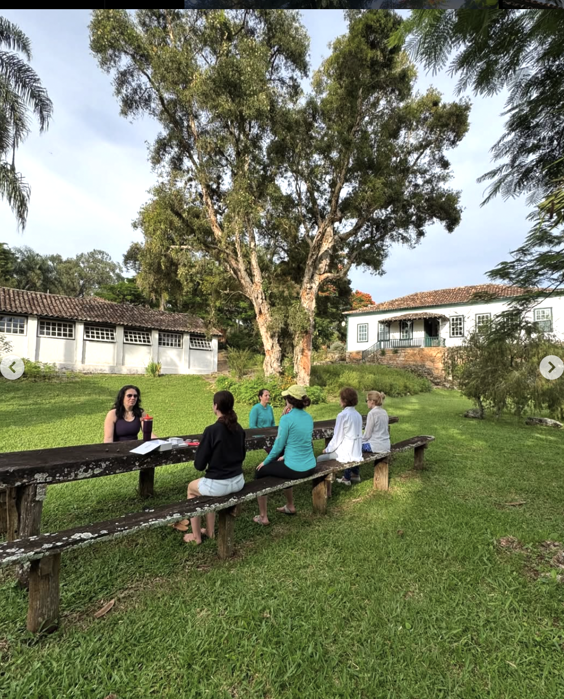 A group of women on retreat sitting on a rustic wooden bench outdoors in front of a large tree, with a white house and lush green grass in the background.
