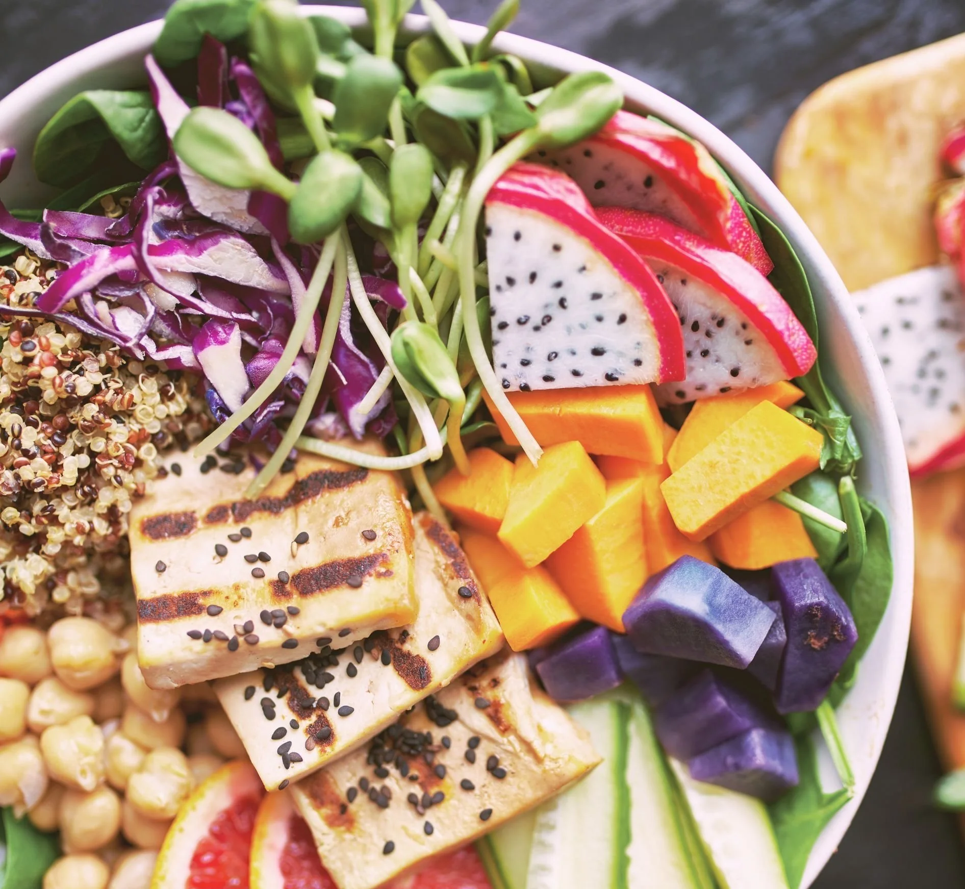 Colorful salad with diced mango, purple sweet potato, sliced dragon fruit, chickpeas, grilled tofu topped with black sesame seeds, shredded purple cabbage, sunflower microgreens, and sliced cucumber.