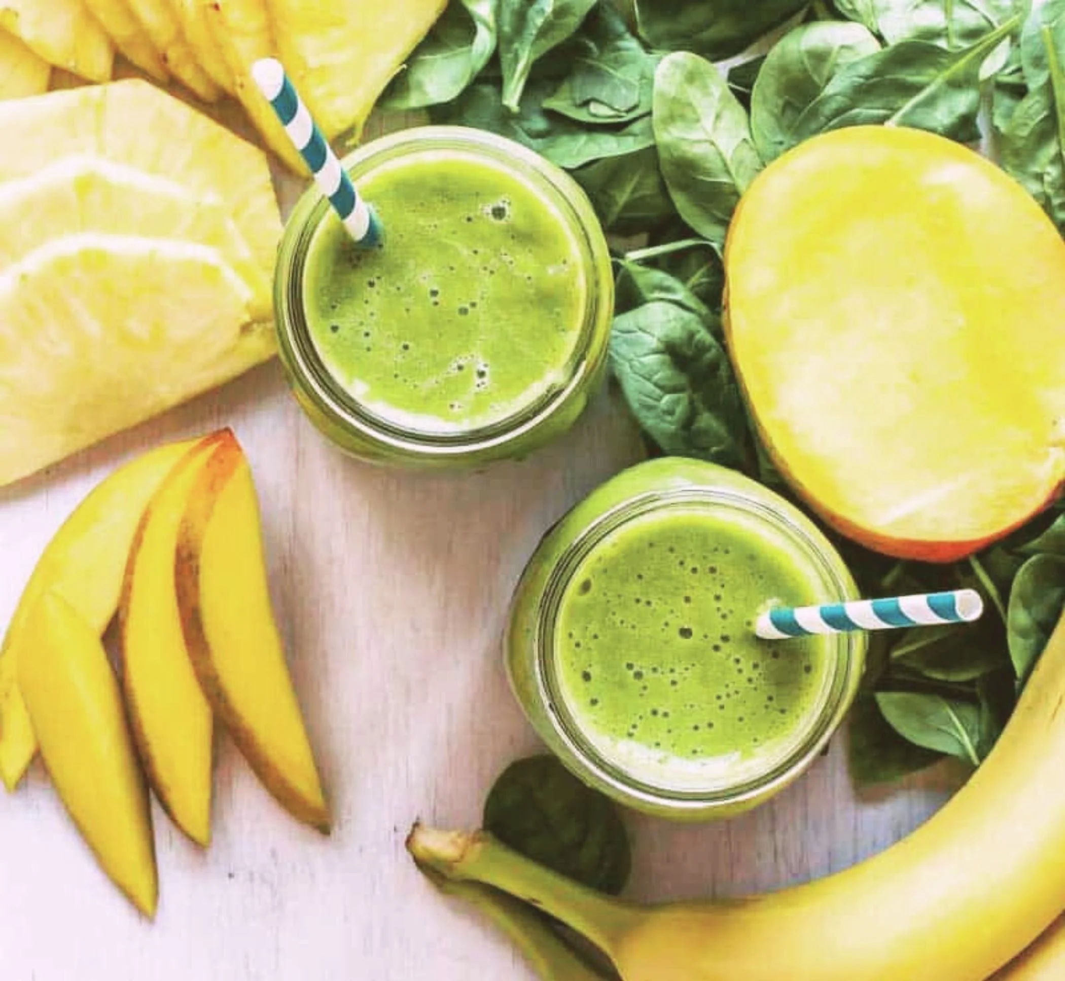 Two glasses of green smoothie on a wooden surface, surrounded by sliced bananas, half a yellow mango, and green leafy vegetables.