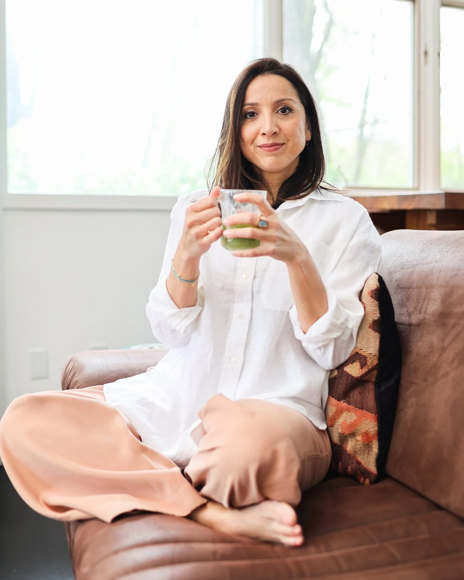 Jessica Sandhu with shoulder-length dark hair, wearing a white shirt and peach-colored pants, sits cross-legged on a brown sofa, holding a glass with a green beverage, in front of a large window with natural light.
