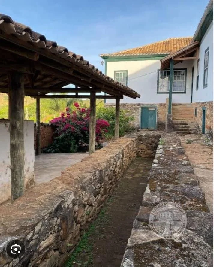 A rustic outdoor scene with a narrow stone-lined irrigation canal running alongside a stone wall. There is a covered porch with wooden posts on the left, and a white house with a green door and windows in the background. Pink flowering bushes are vis