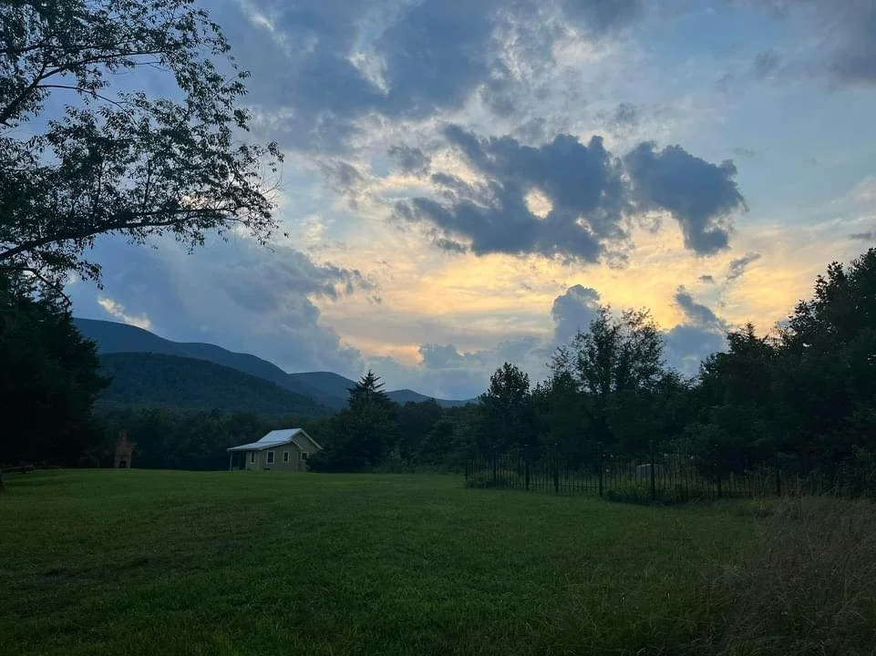 A landscape view at dusk with a grassy field, trees, mountains in the background, and a partly cloudy sky with the sun setting.