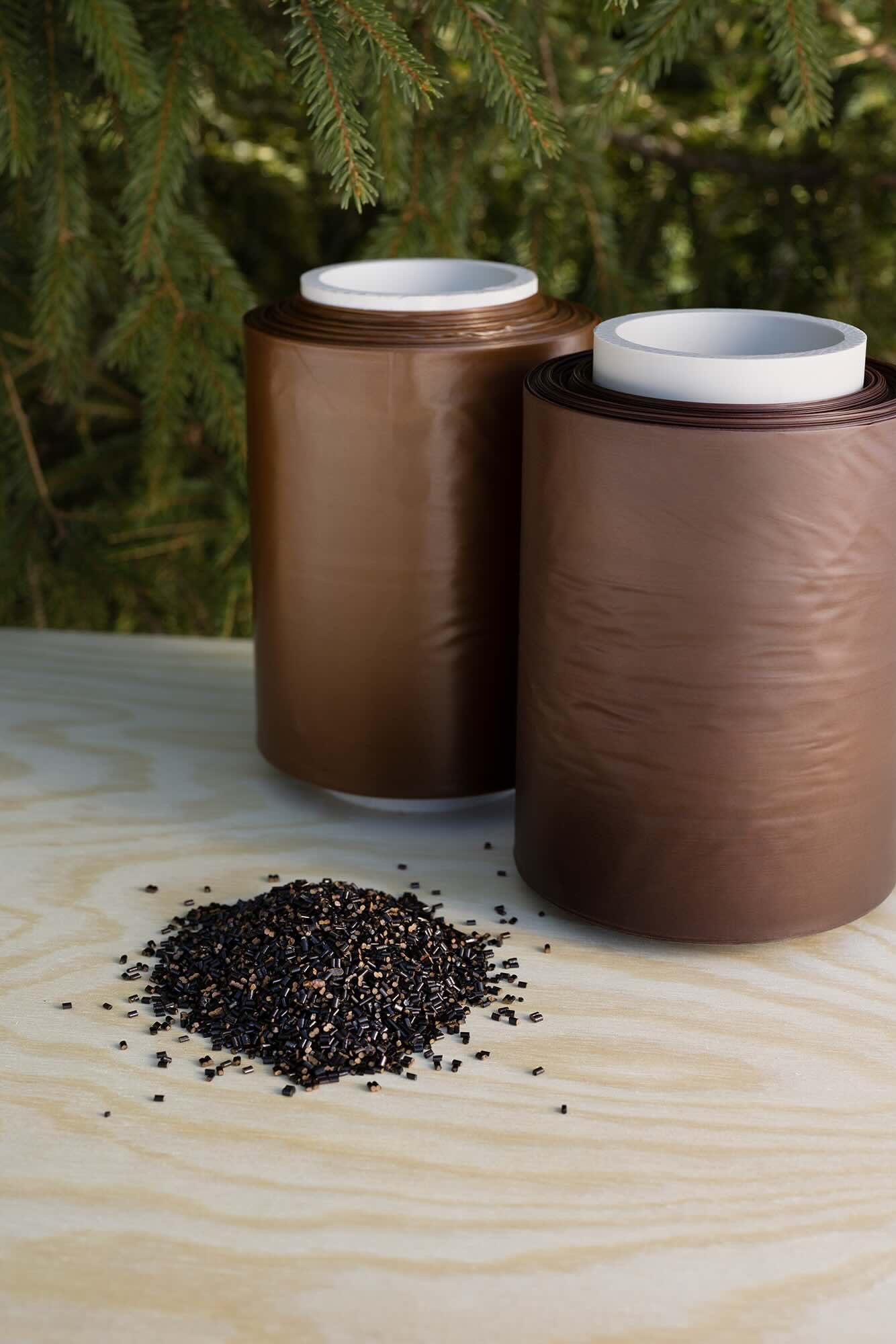 Two rolls of brown polyethylene film and a pile of raw material (Renol pellets) on a wooden table, with spruce branches in the background. Lignin Industries.