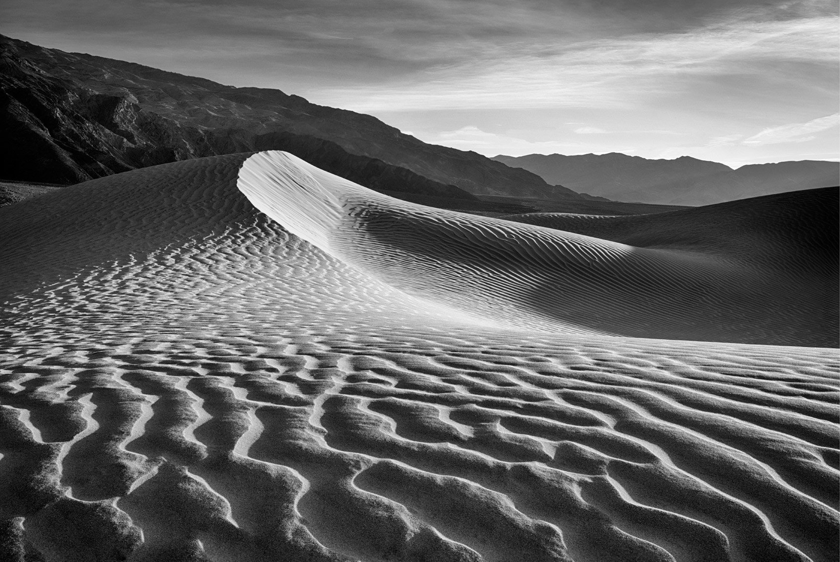 Black and white photograph of ripples of dessert sand with mountains in background