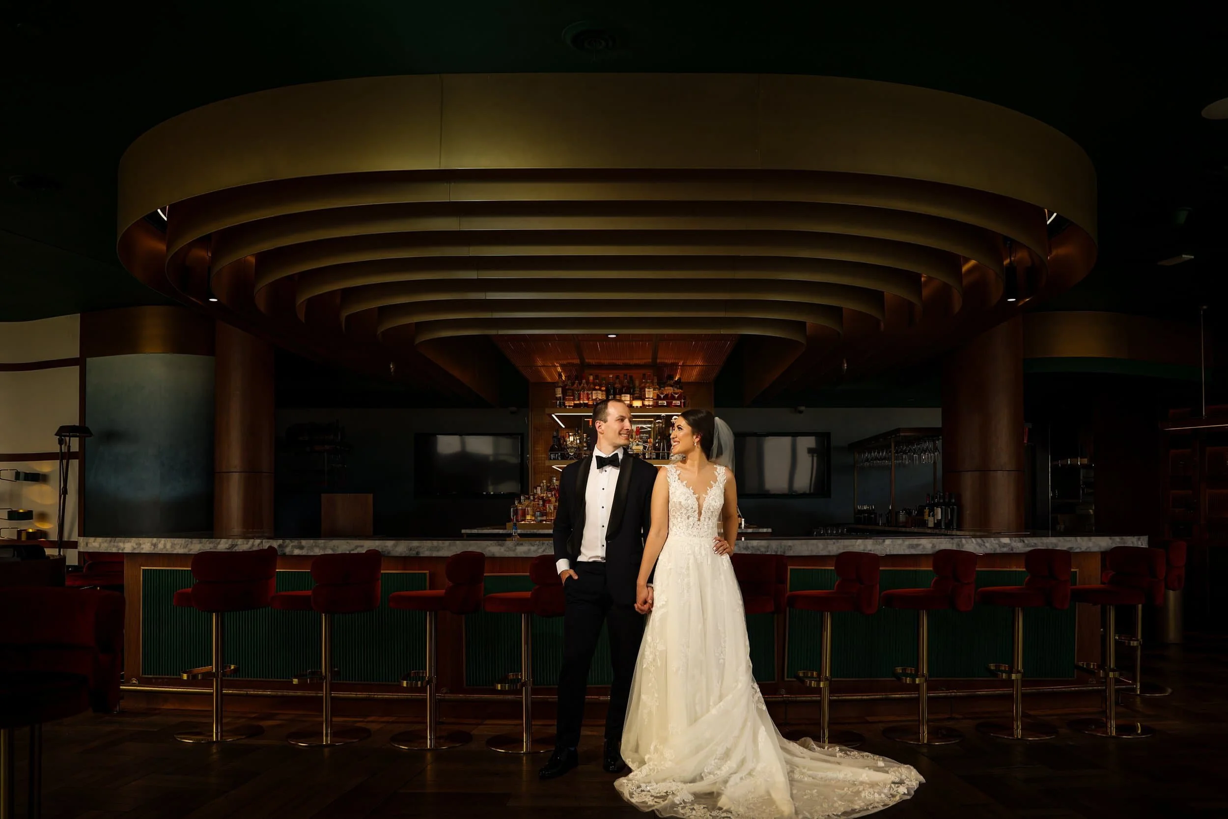 A bride and groom in formal wedding attire stand holding hands in front of an elegant, modern bar with red velvet stools.
