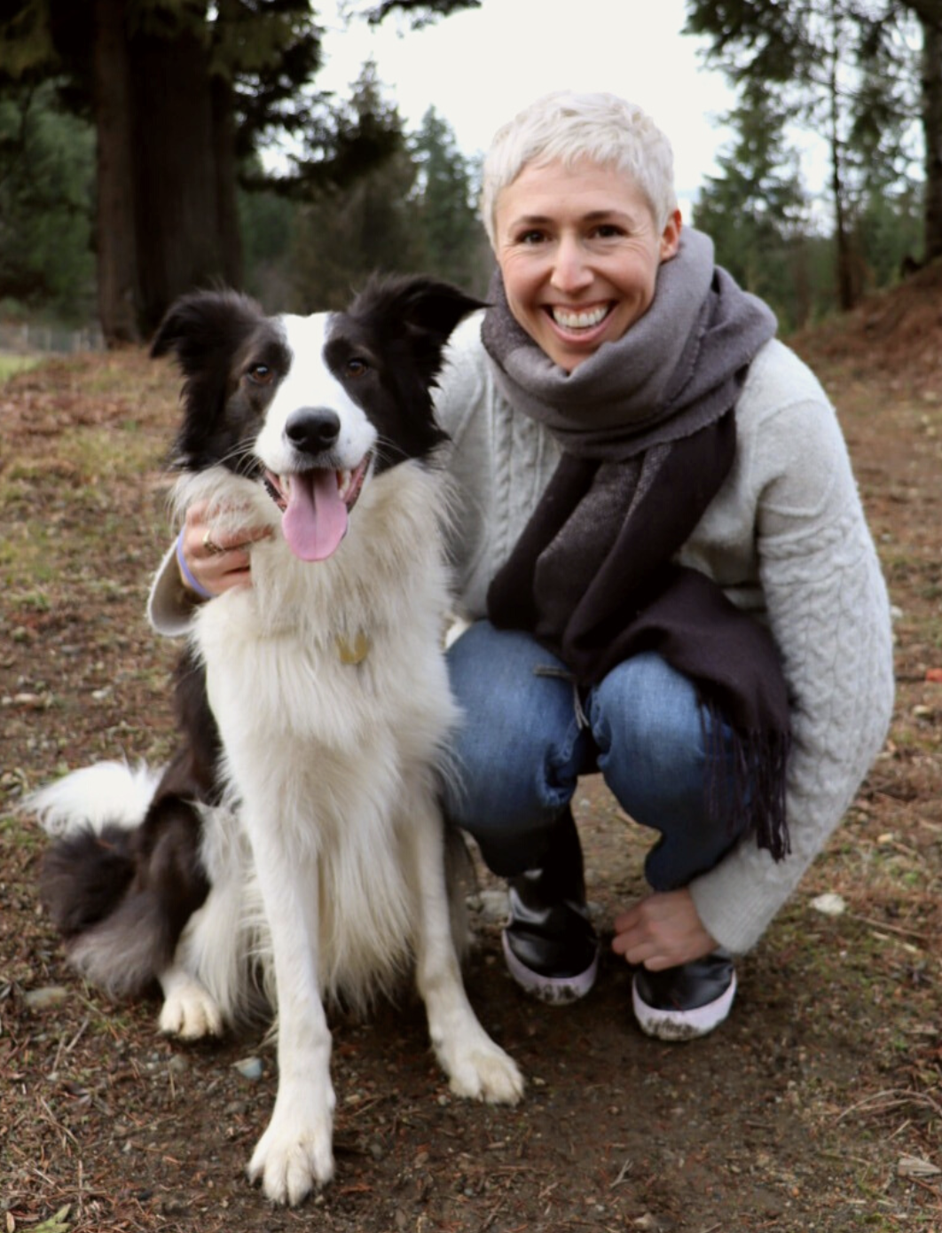 Photograph of female presenting person smiling with short, white hair wearing a scarf, white sweater, jeans and sneakers crouched beside a sitting fluffy black & white dog set against a forest background