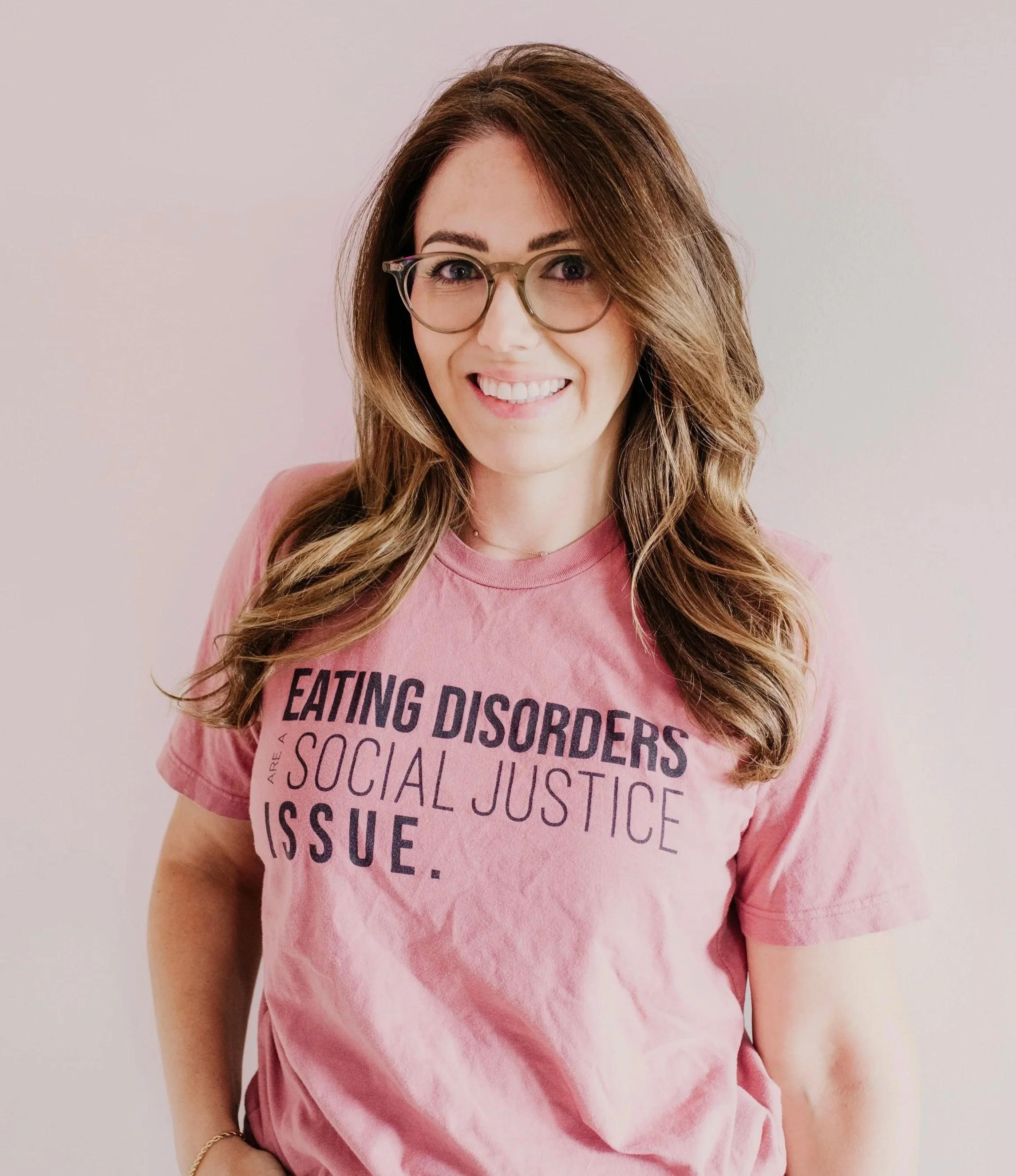 Photograph of a female presenting person smiling with long brown hair, brown glasses and a pink t-shirt with the words, "Eating Disorders are a Social Justice Issue" set against a neutral background