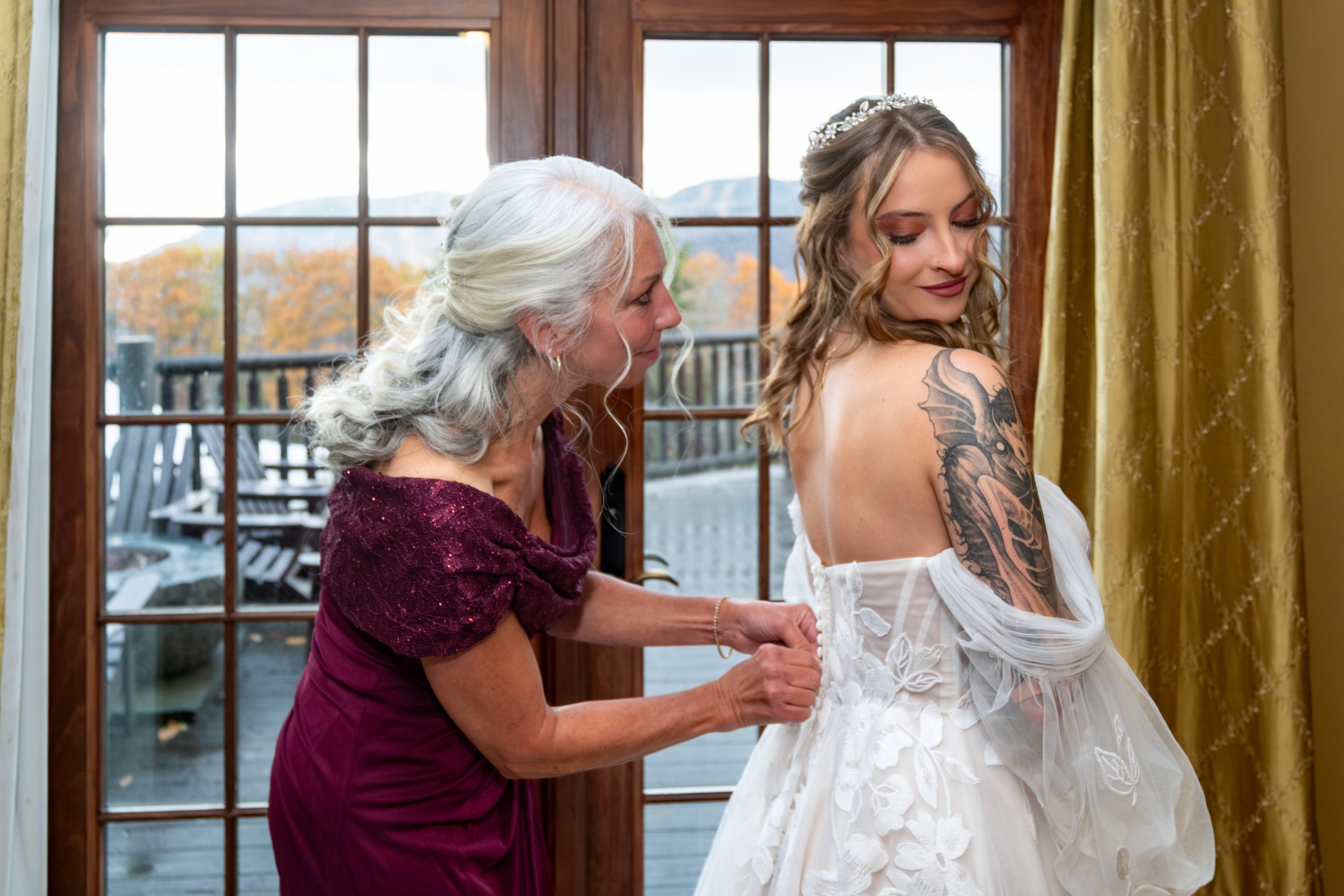 A bride in a lace gown getting ready beside a large window with mountains and fall foliage behind her