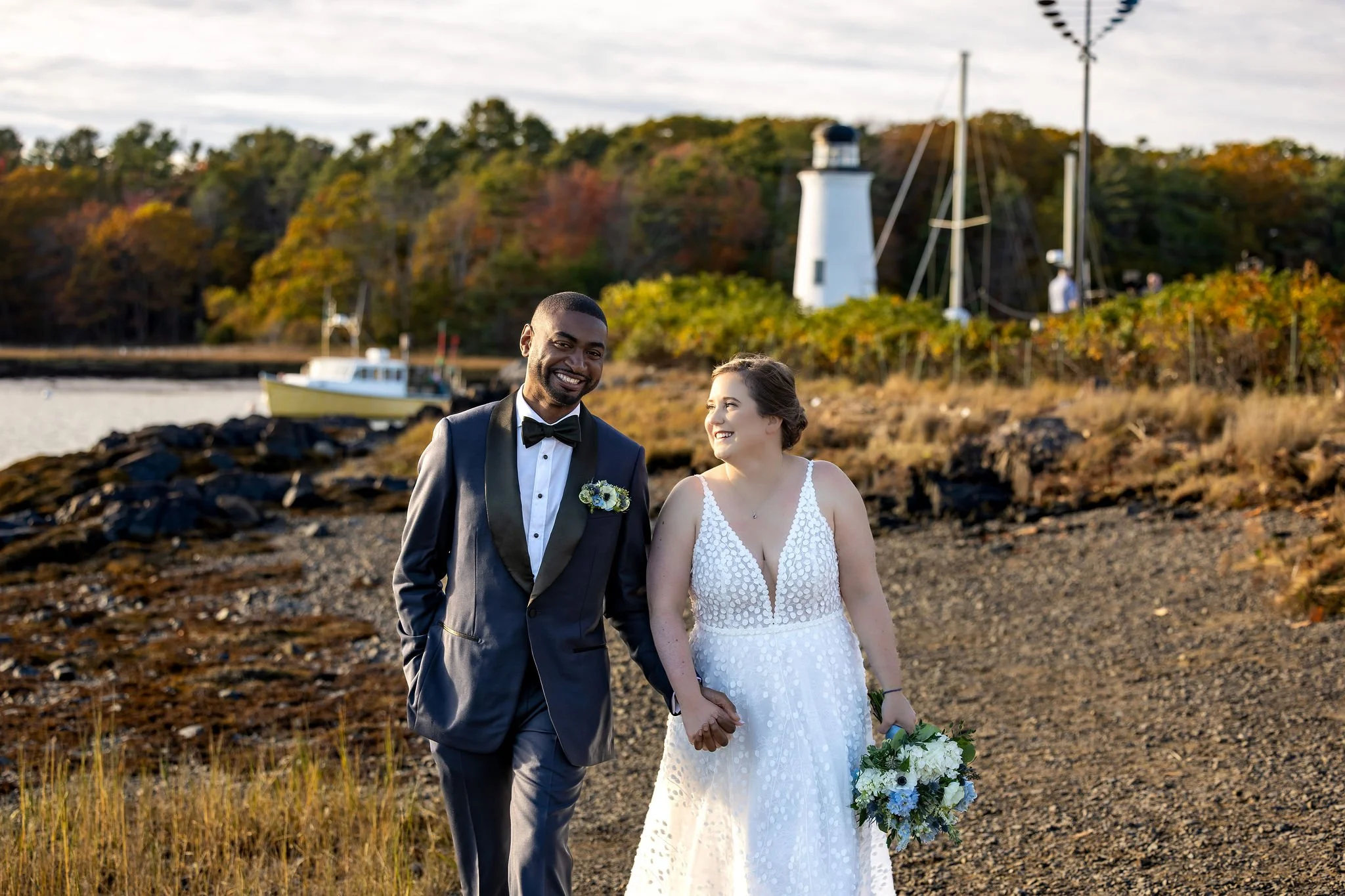 Kaitlyn and Darren walking hand in hand on rocky shoreline smiling Kennebunkport Maine fall wedding portraits VMG Productions