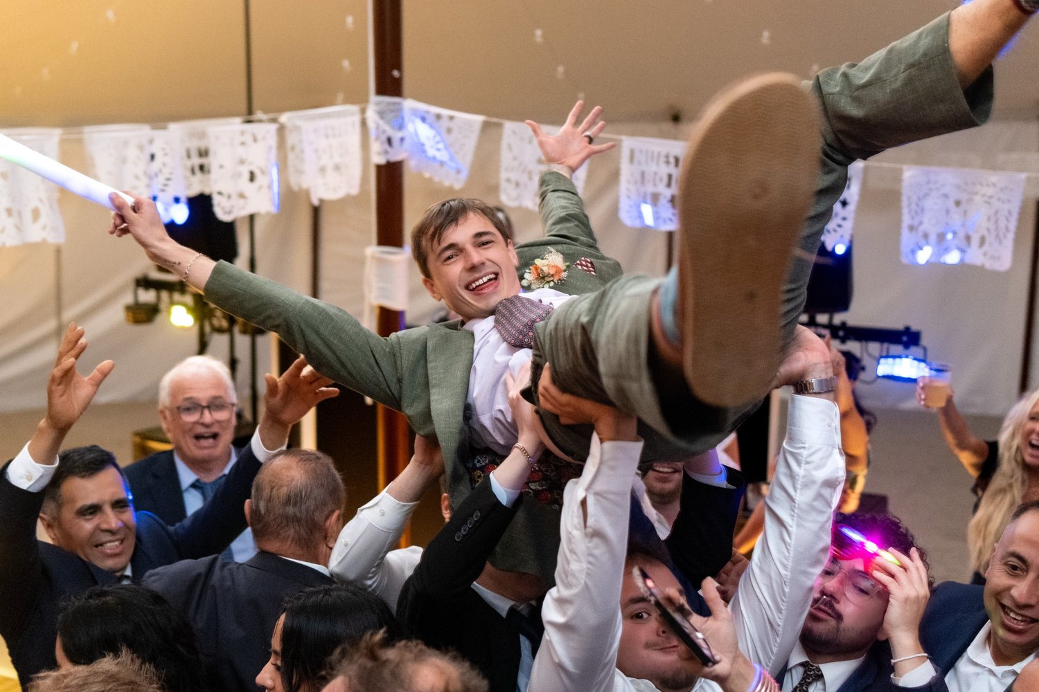 Groom lifted by guests under papel picado banners at tented reception, Wilkinson wedding, Maine