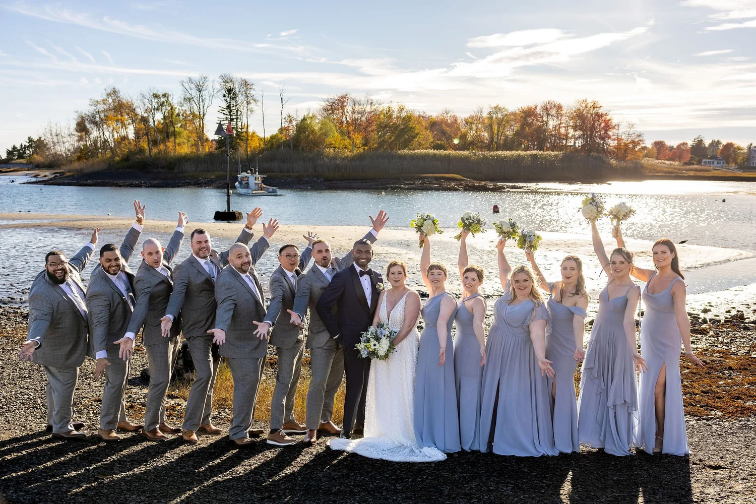 Full wedding party arms raised celebrating on rocky Maine shoreline Kennebunkport fall wedding VMG Productions