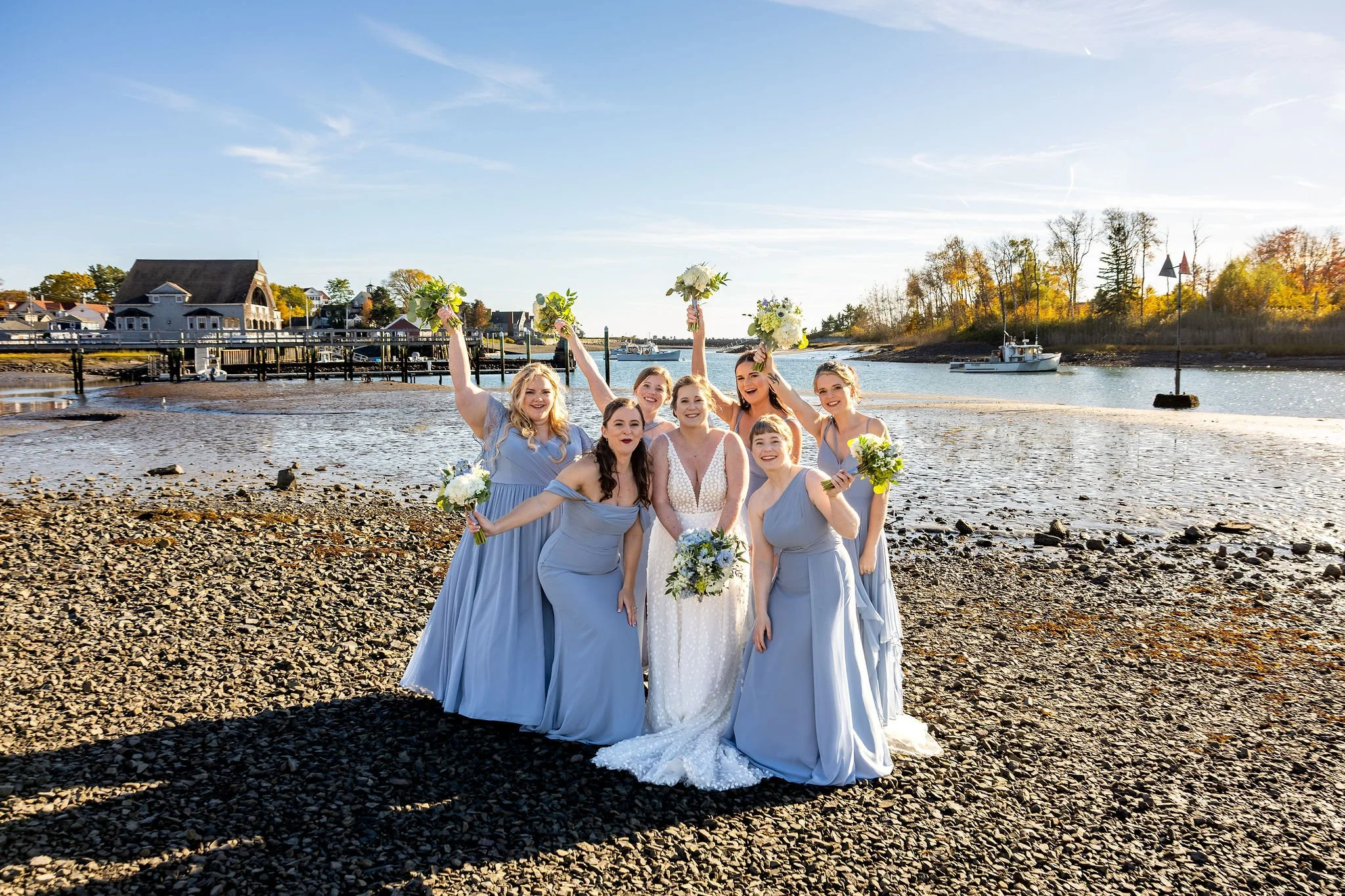 Kaitlyn with bridesmaids celebrating with bouquets raised on rocky shoreline Kennebunkport Maine fall wedding VMG Productions